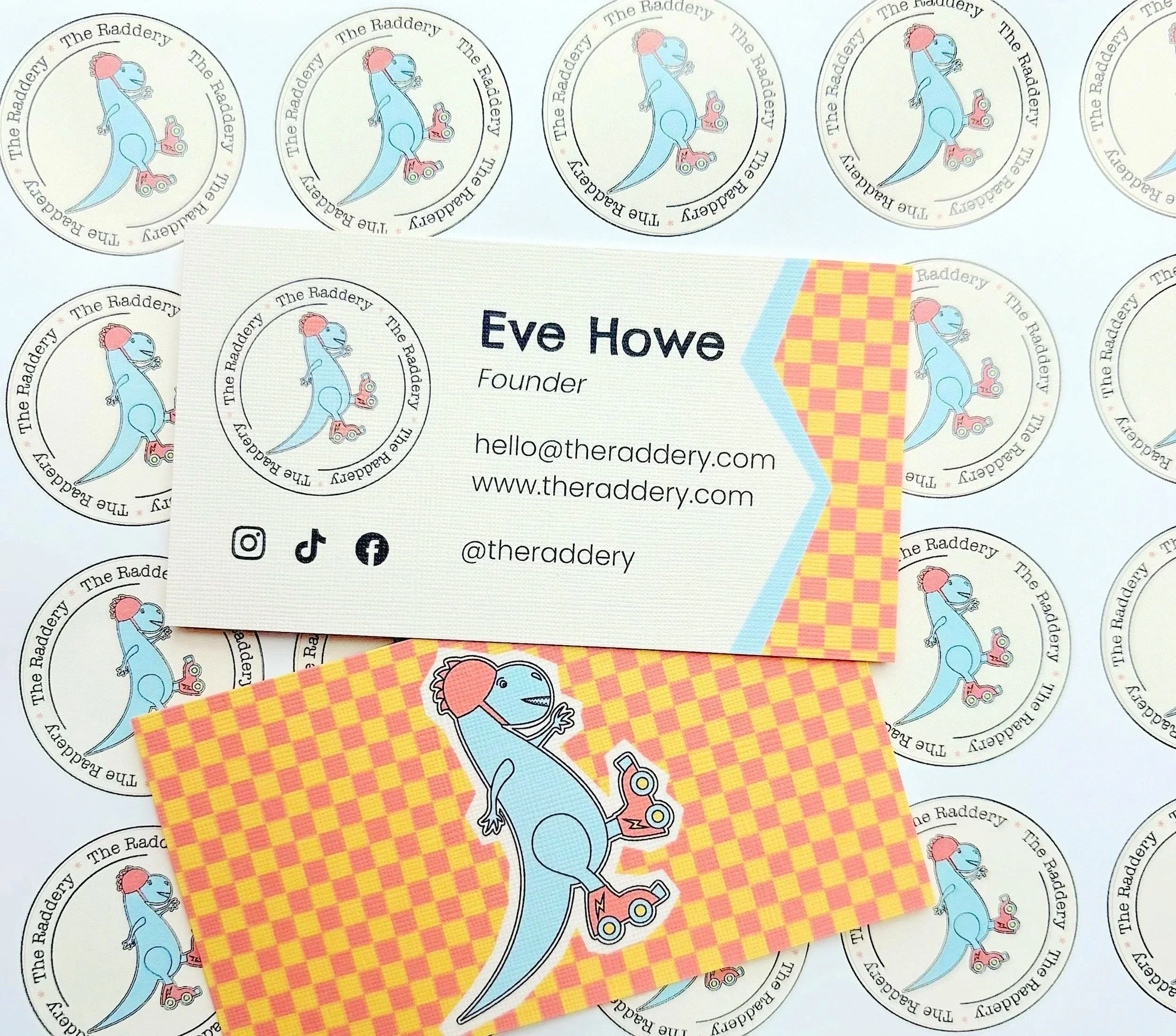 Business cards for 'The Raddery' featuring a blue dinosaur mascot with roller skates and a red hairdo, displayed on a surface with round stickers of the same mascot pattern.