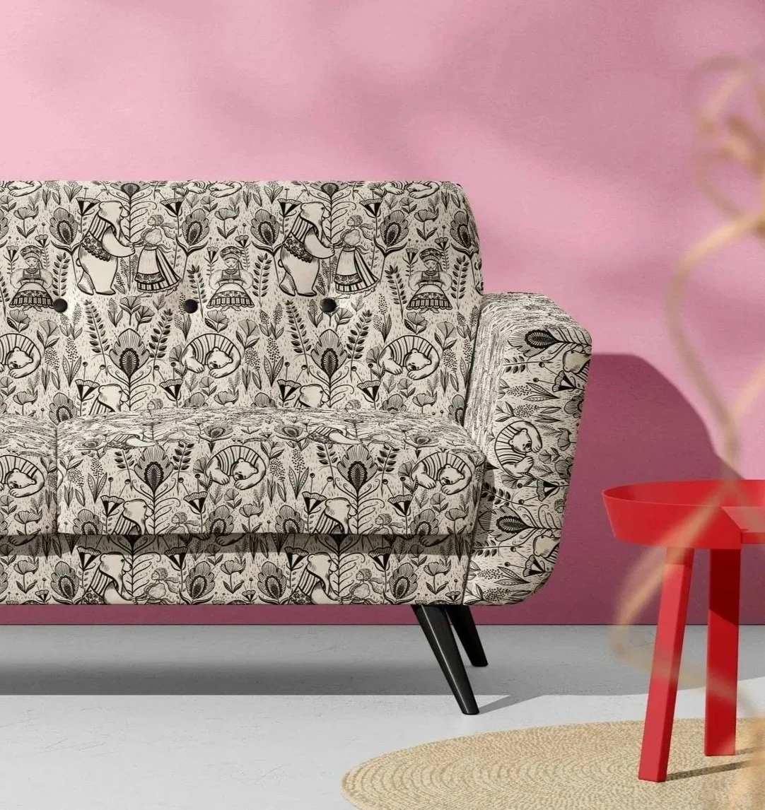 A black and white patterned sofa with animal and floral motifs, positioned against a pink wall, with a red side table and a beige circular rug nearby.