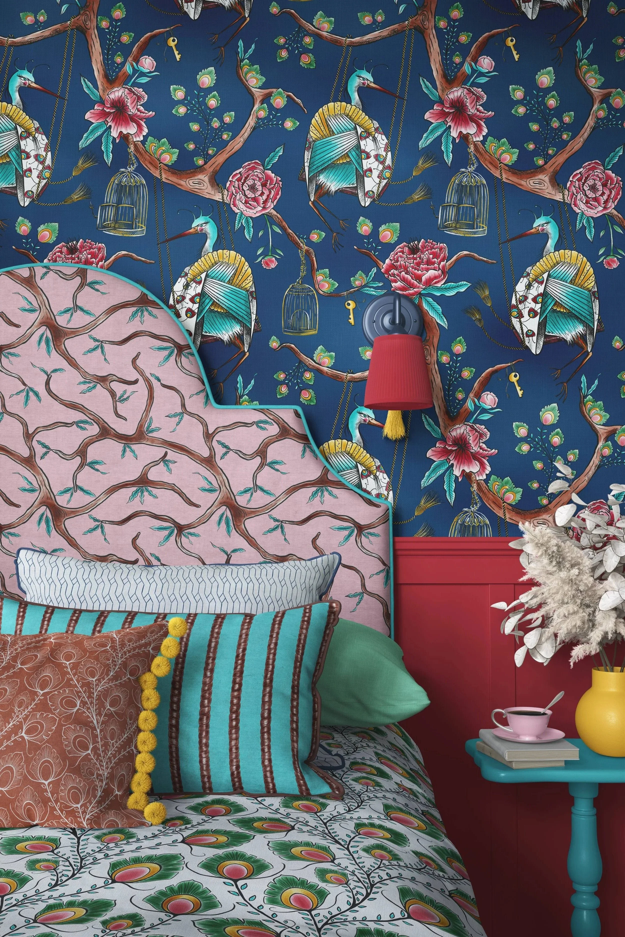 Colorful bedroom with patterned wallpaper featuring bees, pink flowers, and leaves. The bed has multiple patterned pillows with floral, striped, and striped designs. A turquoise side table holds a pink cup, a yellow vase, and books. A red wall-mounted reading light is above the table, and a white fluffy plant is positioned next to it.