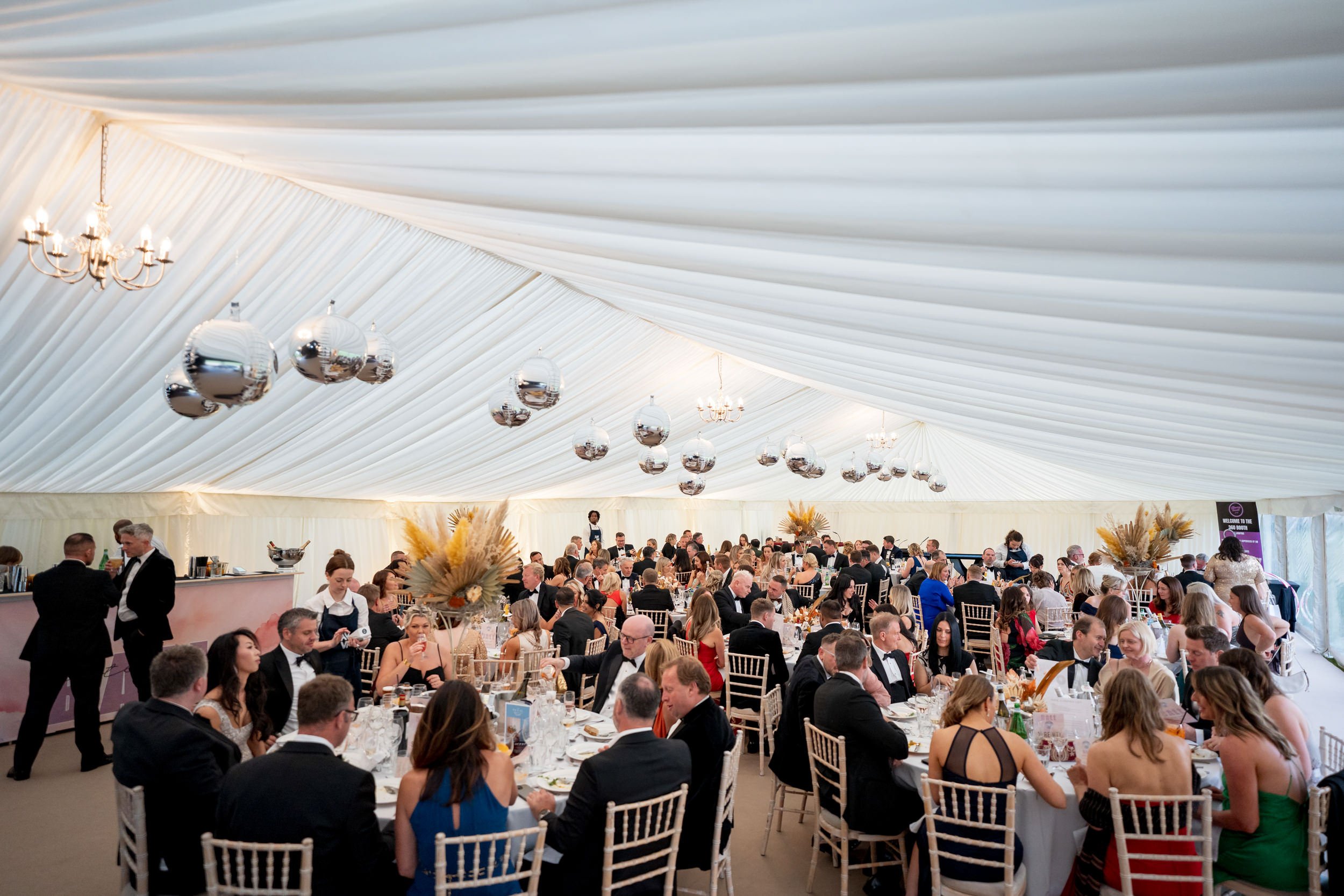 A large formal event or celebration in a spacious tent with many round tables filled with guests dressed in formal attire. Elegant floral arrangements and hanging silver disco balls decorate the space. Attendees are eating, chatting, and enjoying the