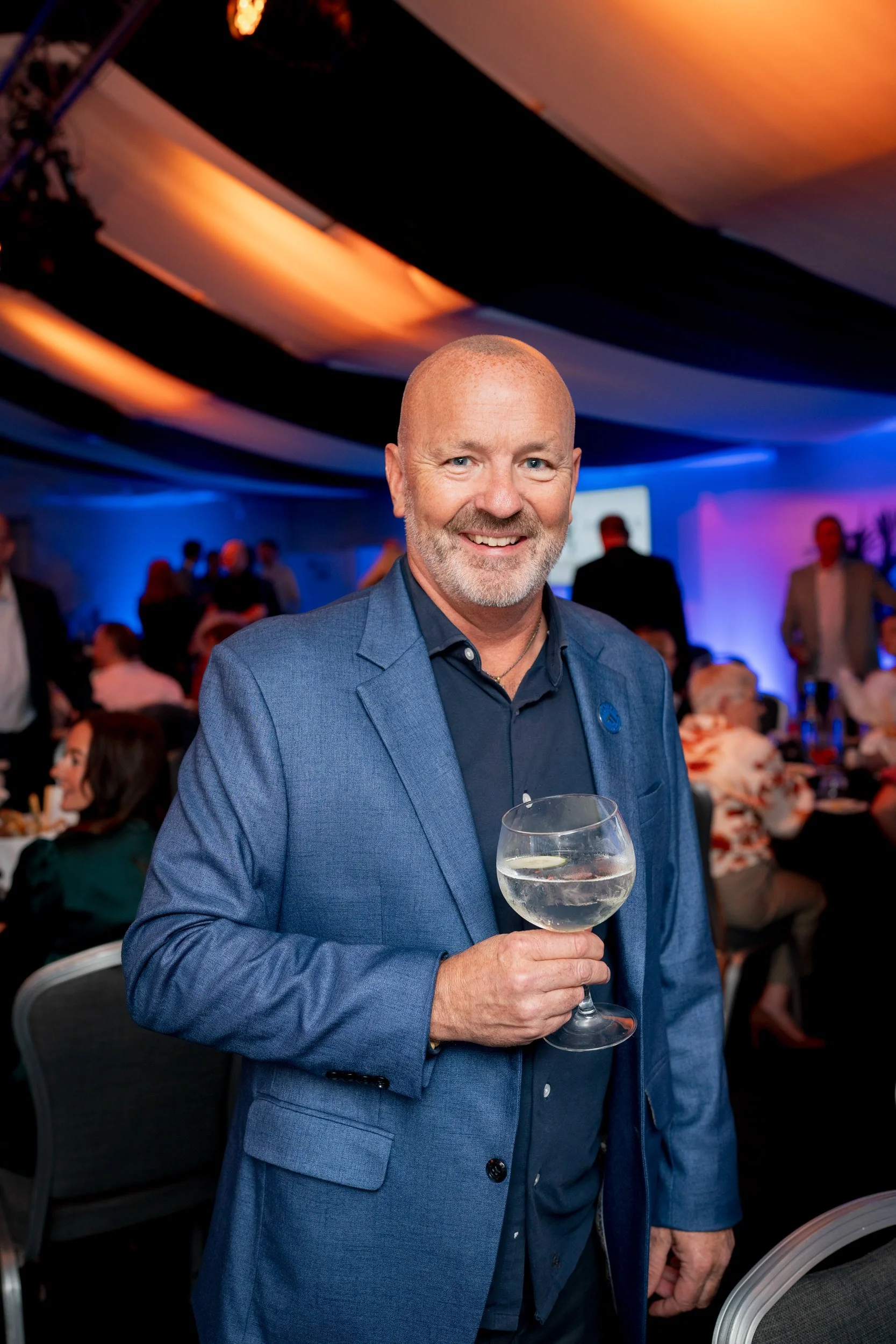 A man with a bald head and gray beard, dressed in a blue suit and navy shirt, holding a glass of water at an evening reception for the NAFD conference event with colorful lighting and people in the background.