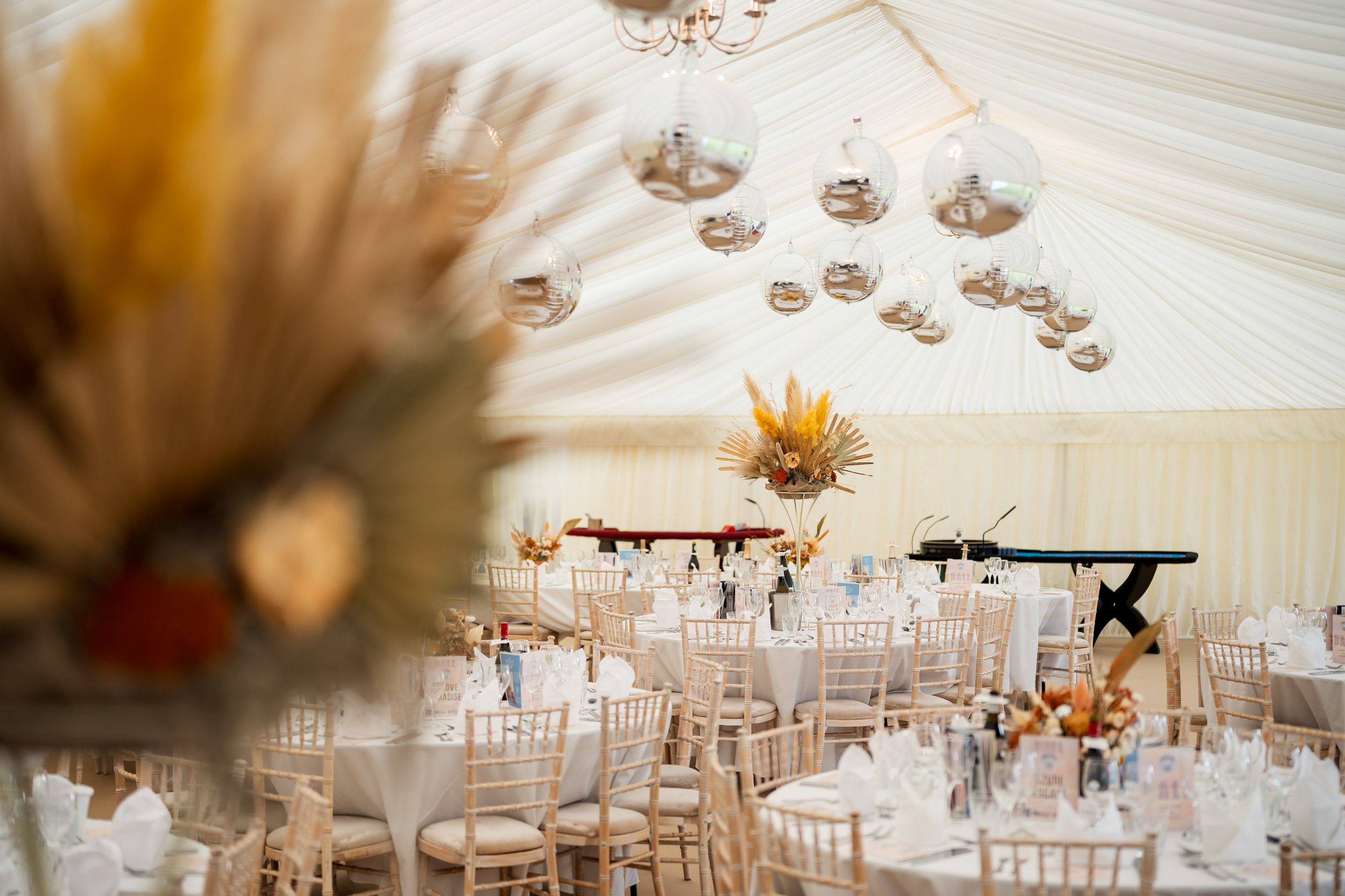 Elegant event tent decorated for a celebration with round tables set with white tablecloths, floral centerpieces, and gold chairs. Hanging glass orb lights are suspended from the ceiling, and a large floral arrangement is in the foreground.