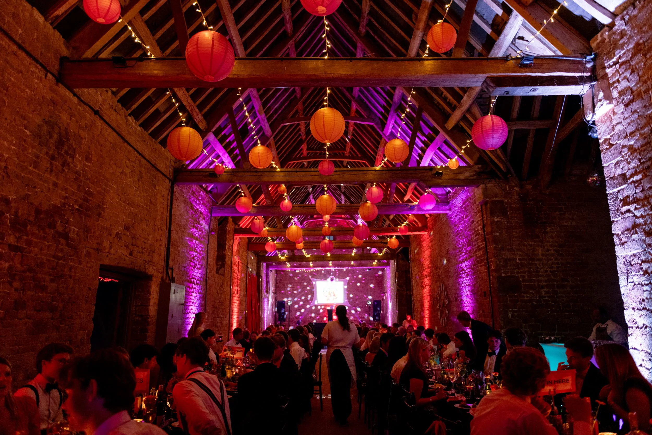 Decorated brick barn walls and a high wooden ceiling, adorned with orange paper lanterns and strings of fairy lights, with purple lighting and a stage at the front where a presentation screen is displaying a birthday message.