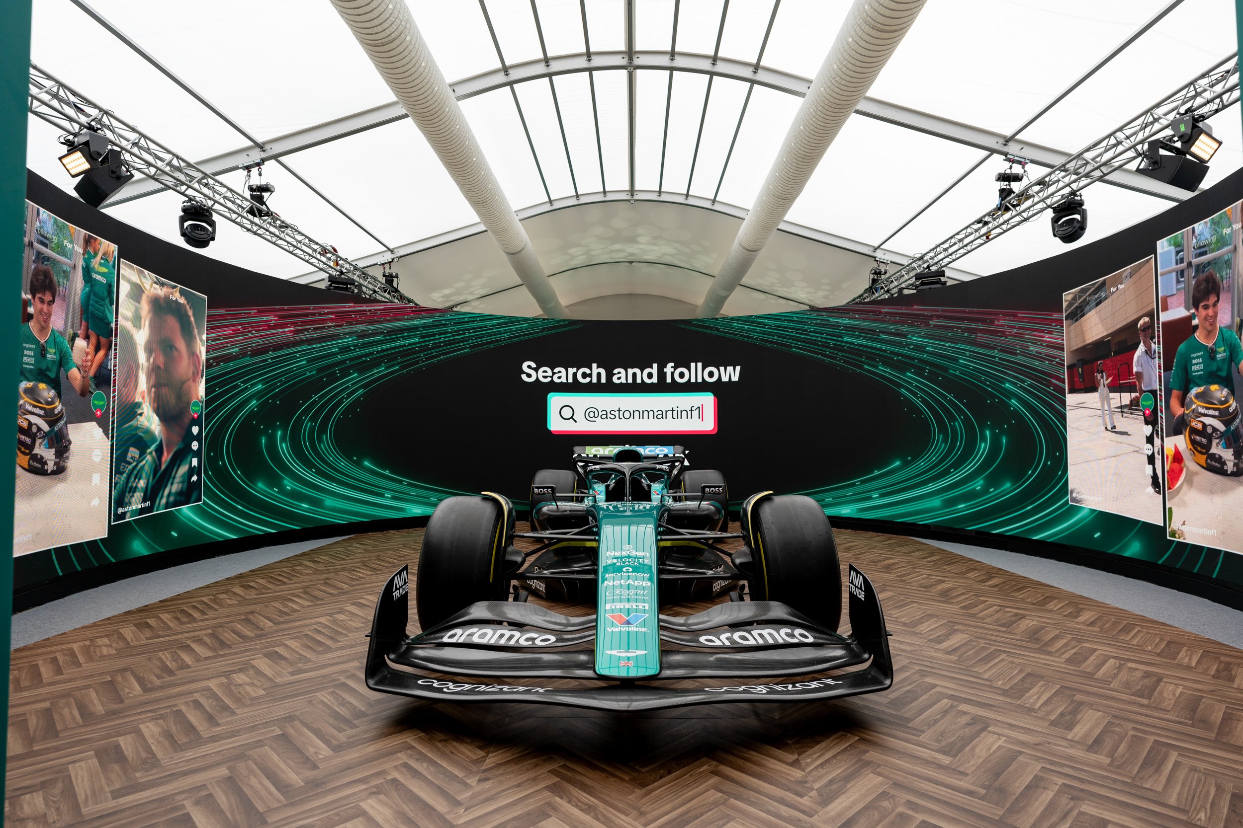 A Formula 1 race car on display in front of a large digital screen with a search prompt to follow an Instagram account, inside a Fews Marquee for Aston Martin Hospitality at Silverstone