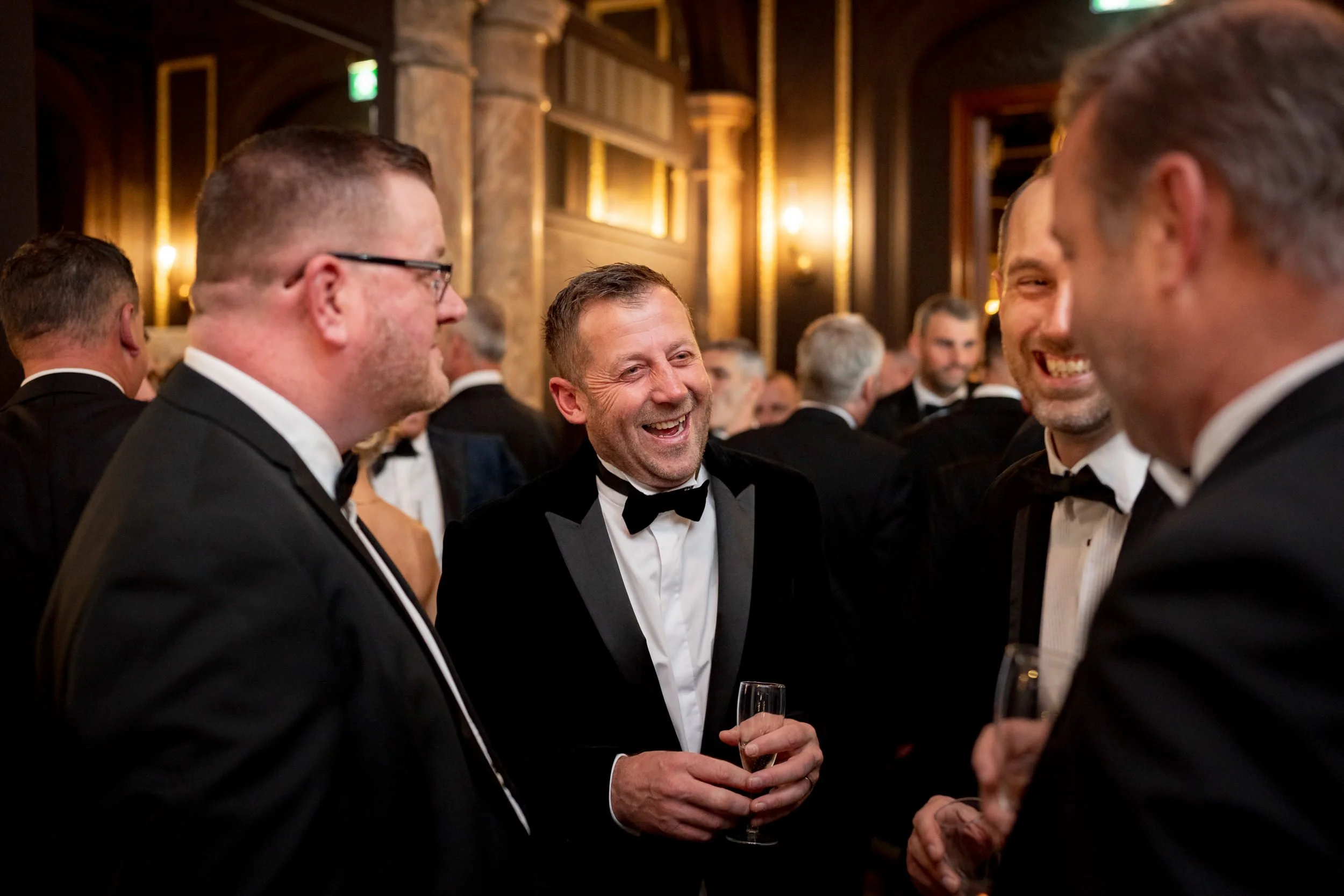 Men in formal tuxedos laughing and talking at an awards evening event, holding champagne glasses, in a lavish, brick and wood interior.