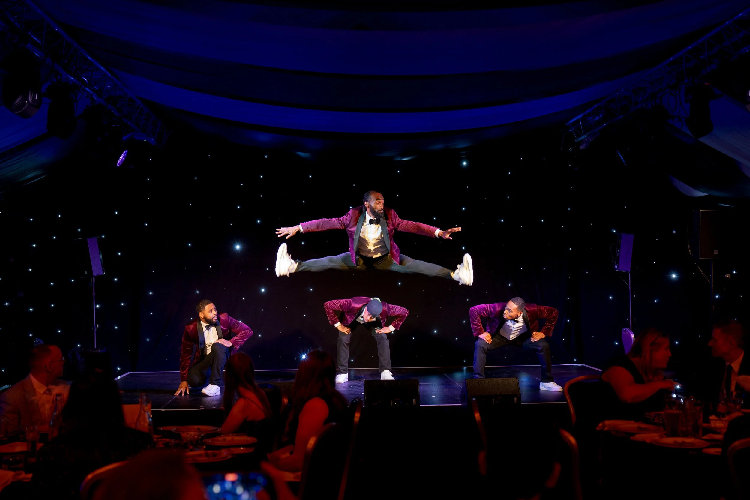 Four male performers in suits and bow ties on stage, with one performing a jump over the others, during a dance act, with an audience visible in the foreground, and a starry black backdrop with stage lighting