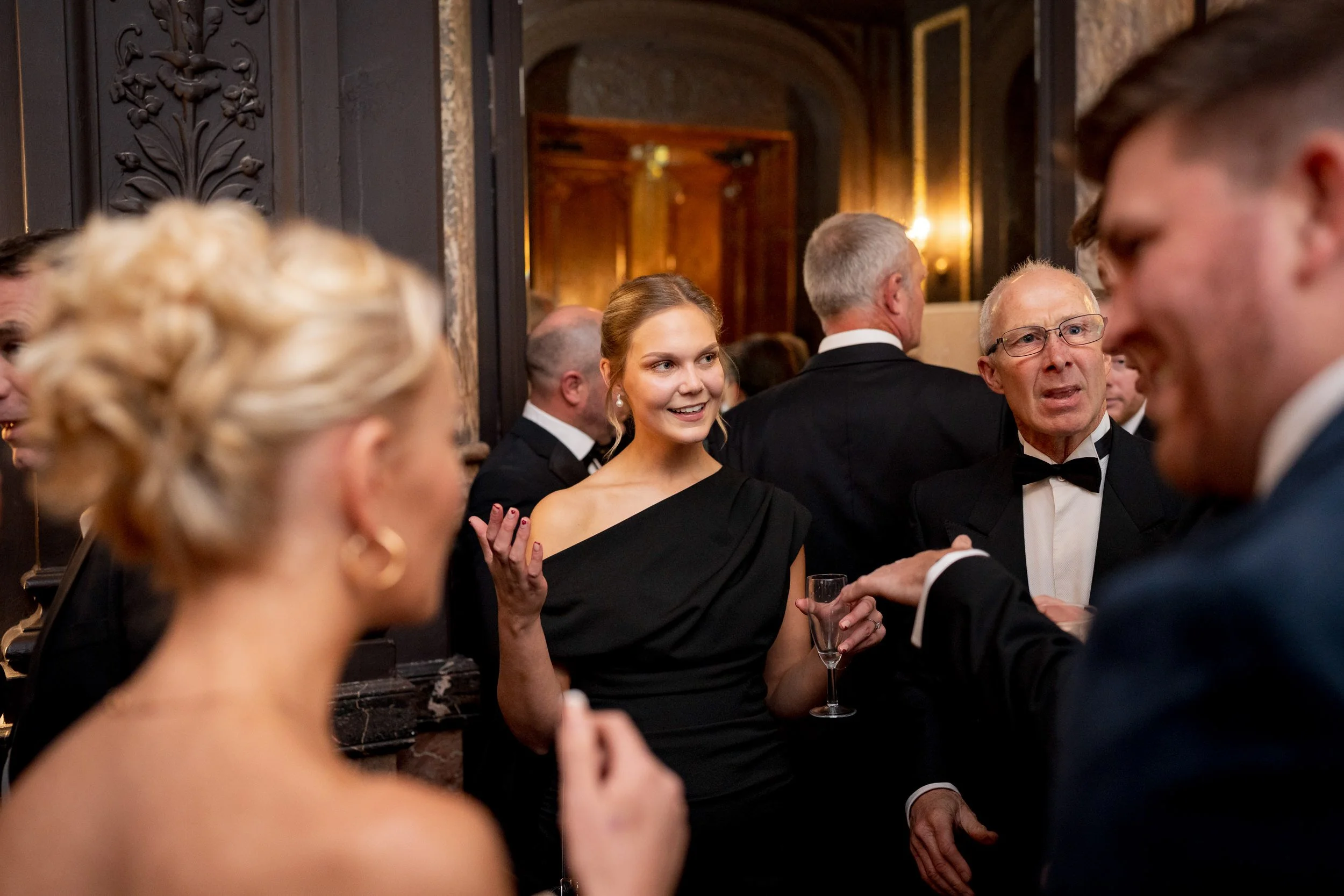 Group of people at a formal awards event, including a young woman in a black off-shoulder dress holding a glass of champagne, talking to older men in tuxedos in an elegant room with dark wood decor.