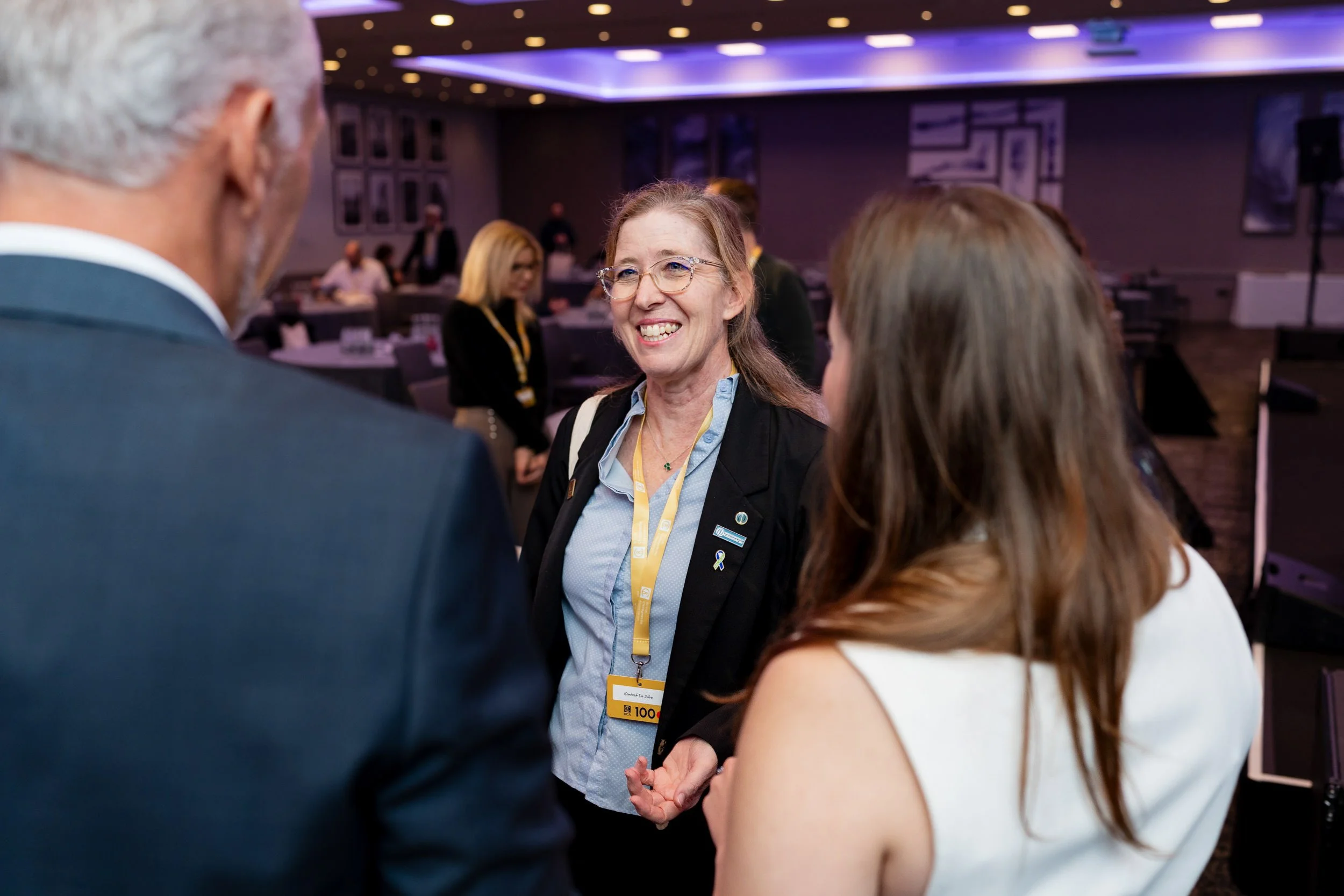 A group of three people in a business conference or networking event, smiling and talking to each other in a well-lit conference room with other attendees in the background.