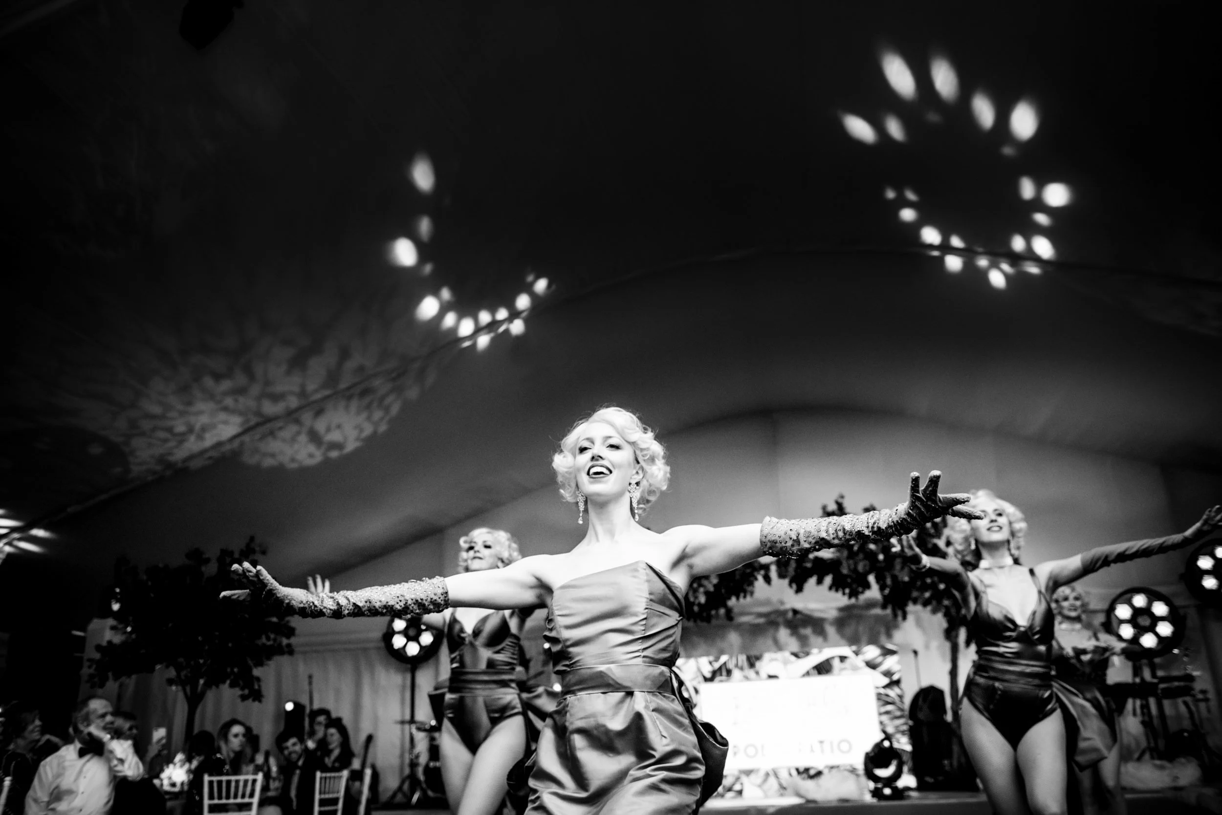 Black and white photograph of women in glamorous 1920s style costumes performing a dance on stage at a luxury party with arms outstretched, smiling, with a decorated backdrop and audience in the background.