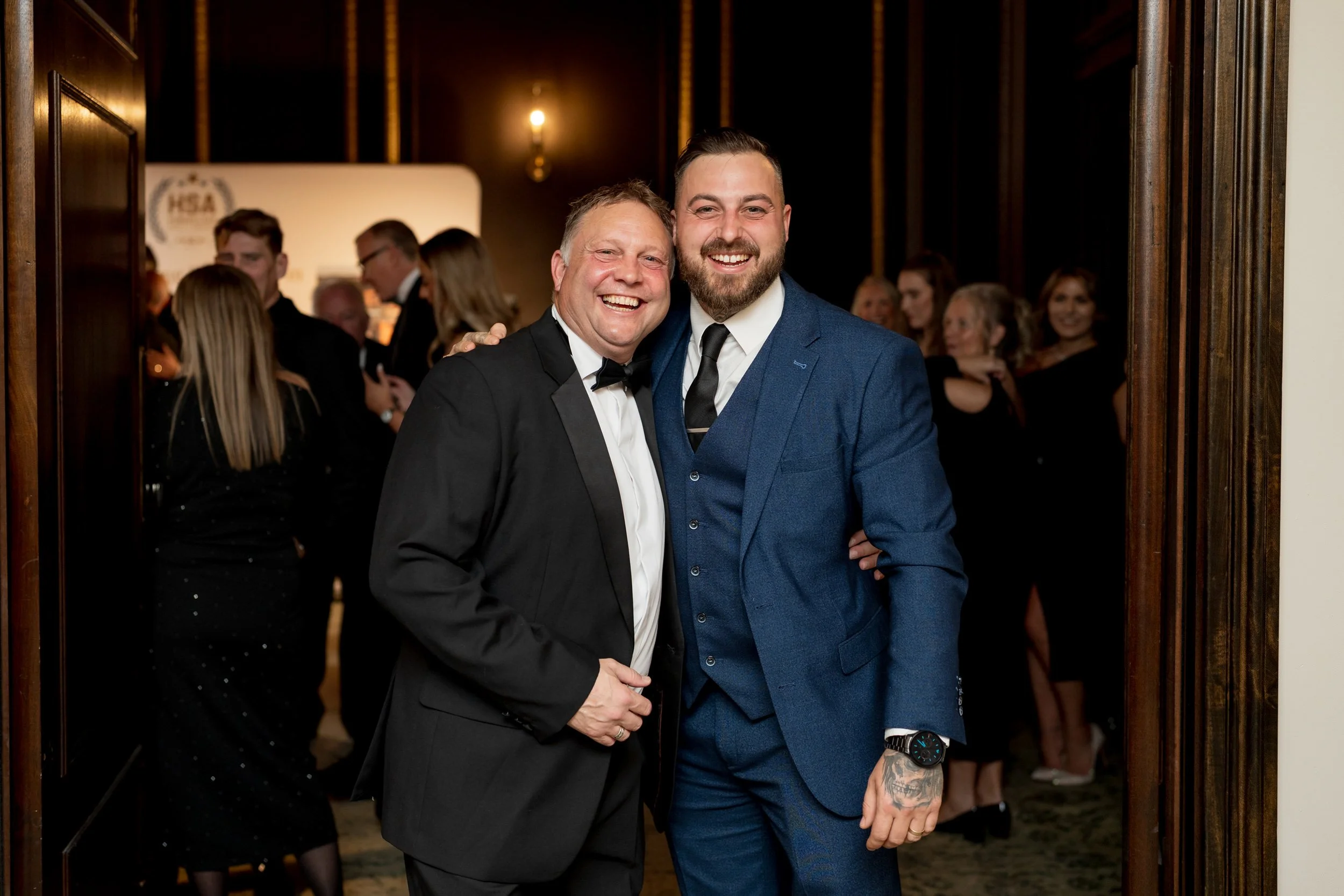 Two men in tuxedos smiling and posing together at an awards formal event, with other guests in the background.