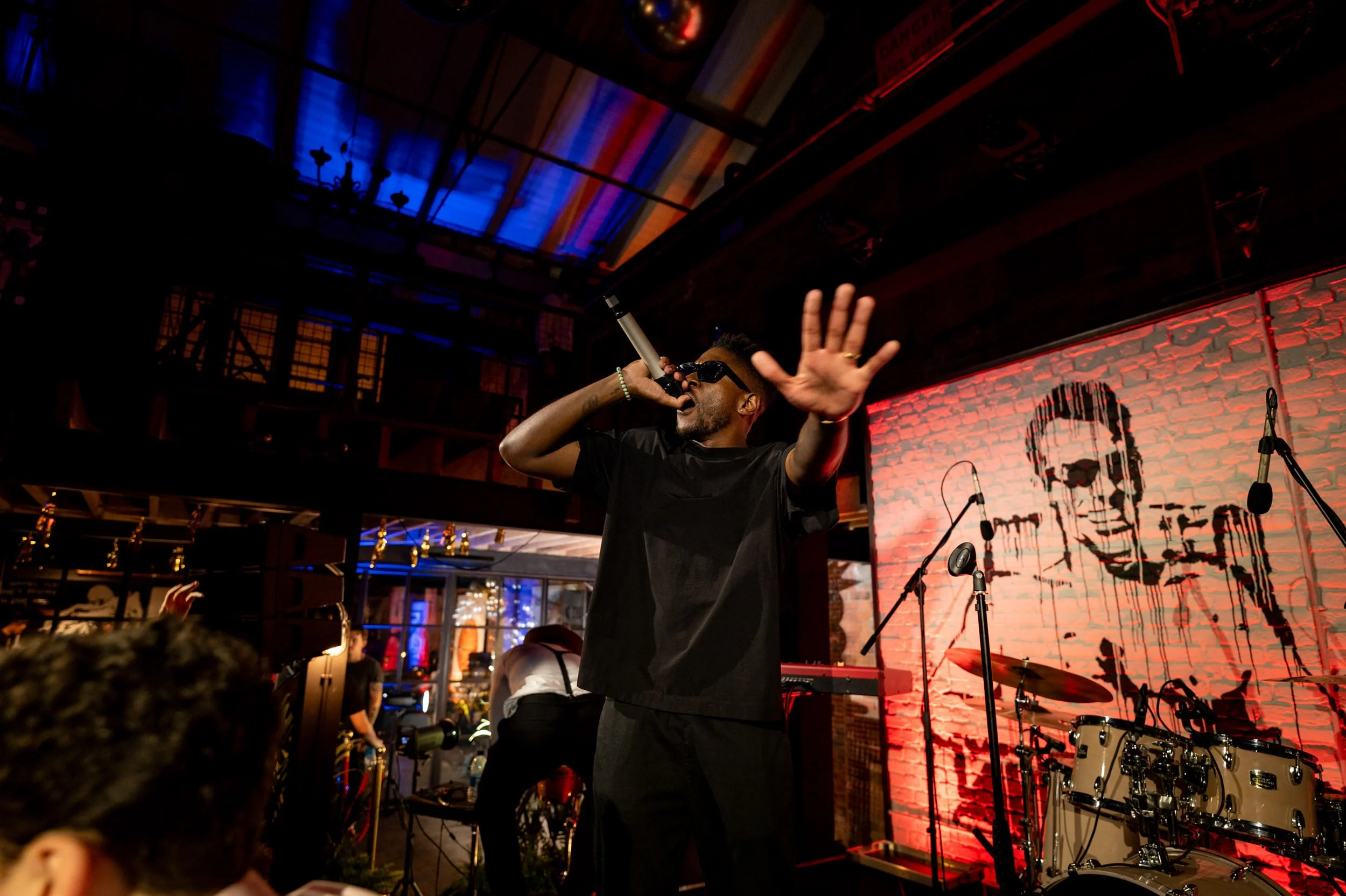 Performer singing into microphone with hand raised, standing on stage with a drummer and abstract portrait mural in background at The Compound in Birmingham for a birthday event