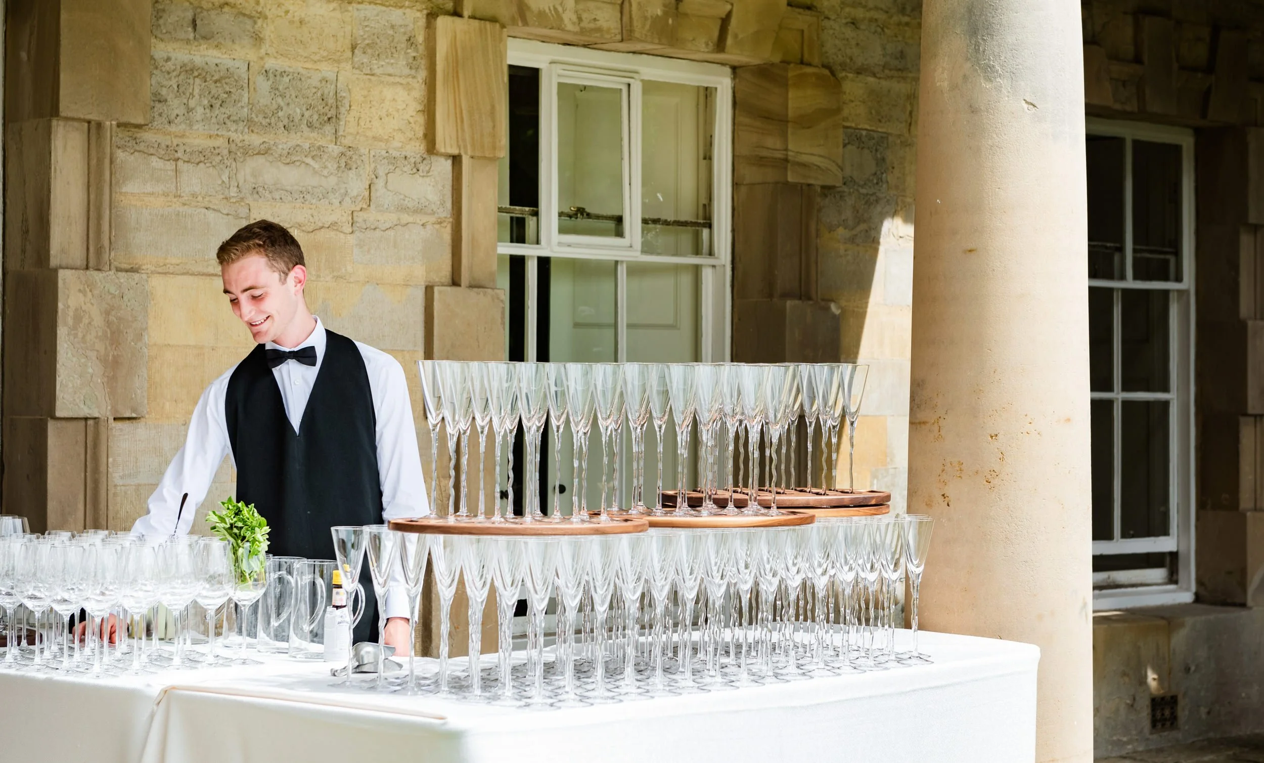 A smiling young male server in a black vest and bow tie stands beside a table set with numerous empty champagne glasses, arranged in tiers on copper trays, at an outdoor event with stone walls and windows in the background.