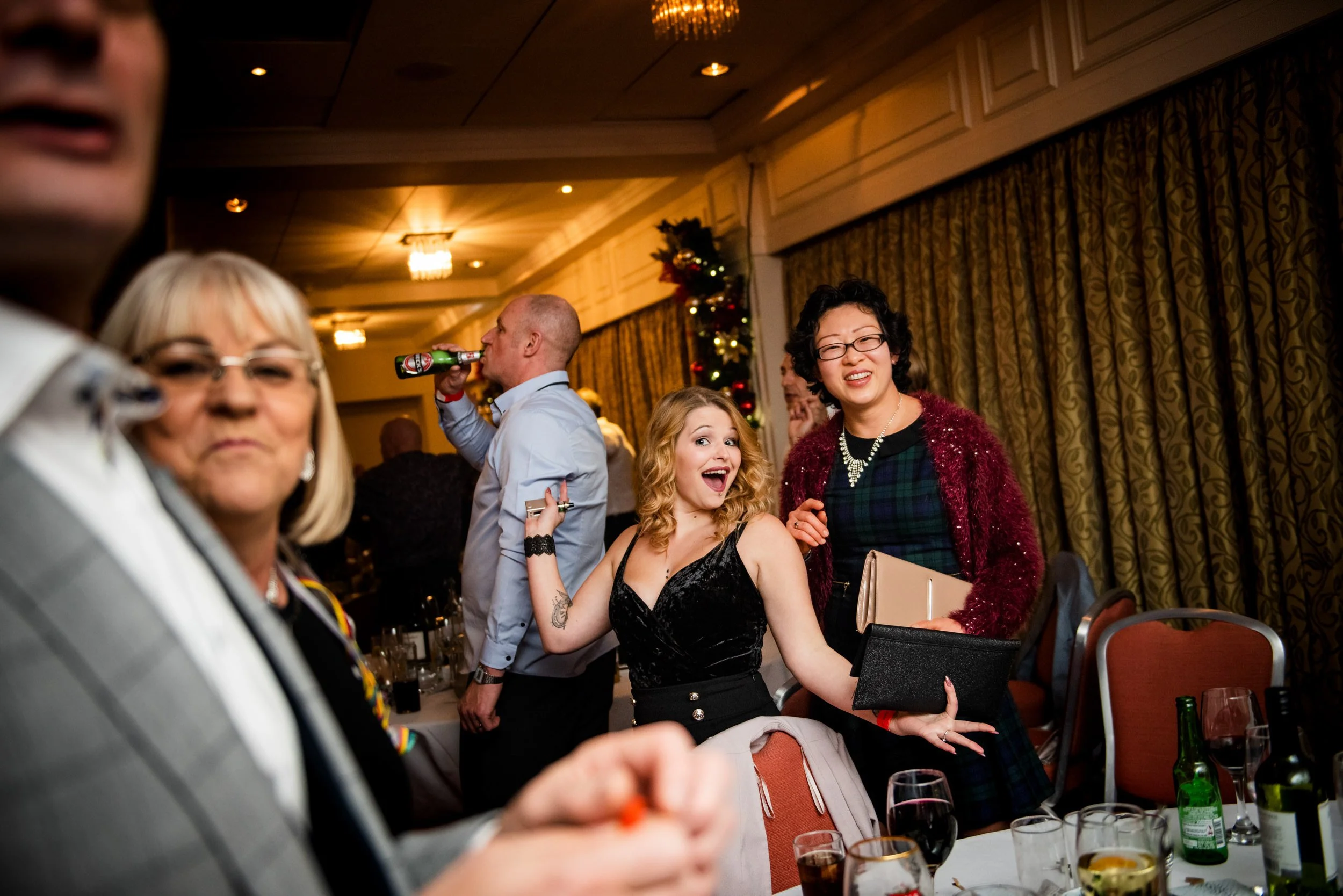 Group of people celebrating at an awards evening, some smiling and others drinking, in a decorated room with Christmas tree, tables, and chairs.