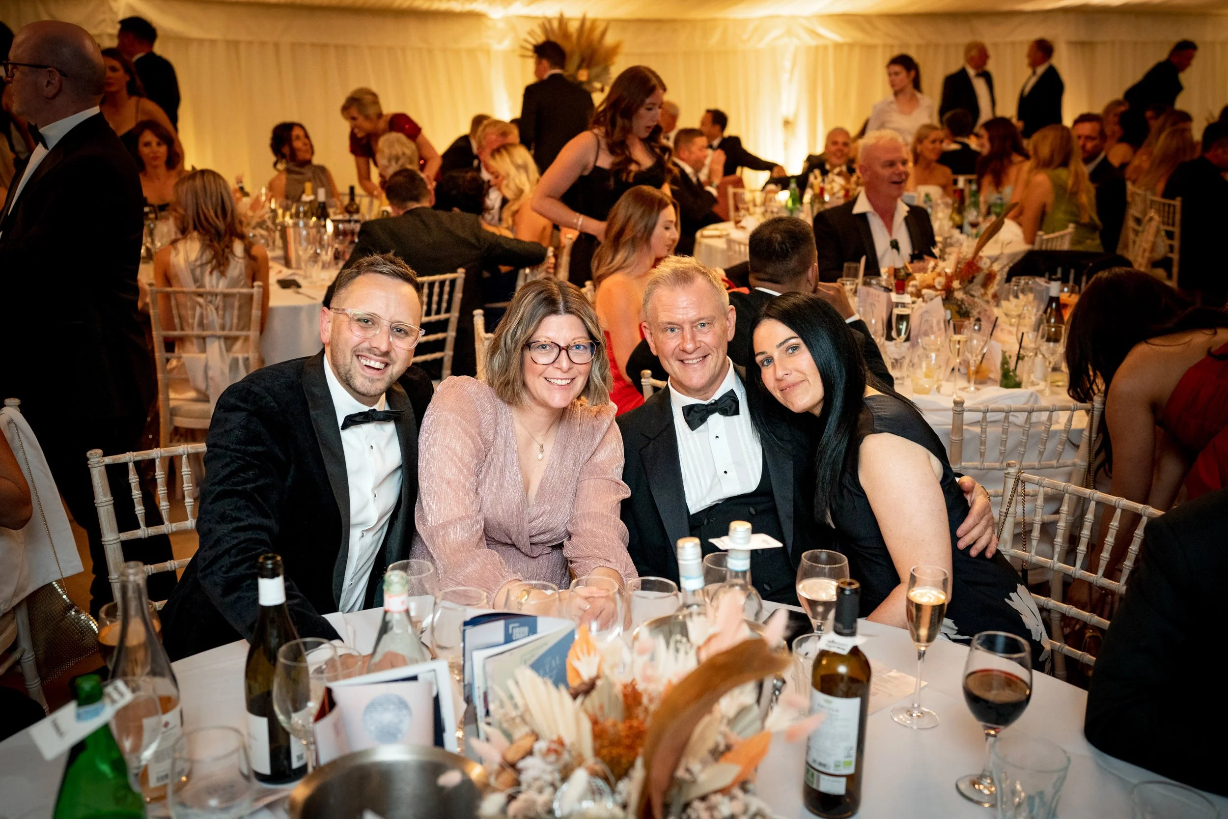 People dressed in formal attire, sitting at a banquet table during a celebration or event, with tables, chairs, and decorations in the background.
