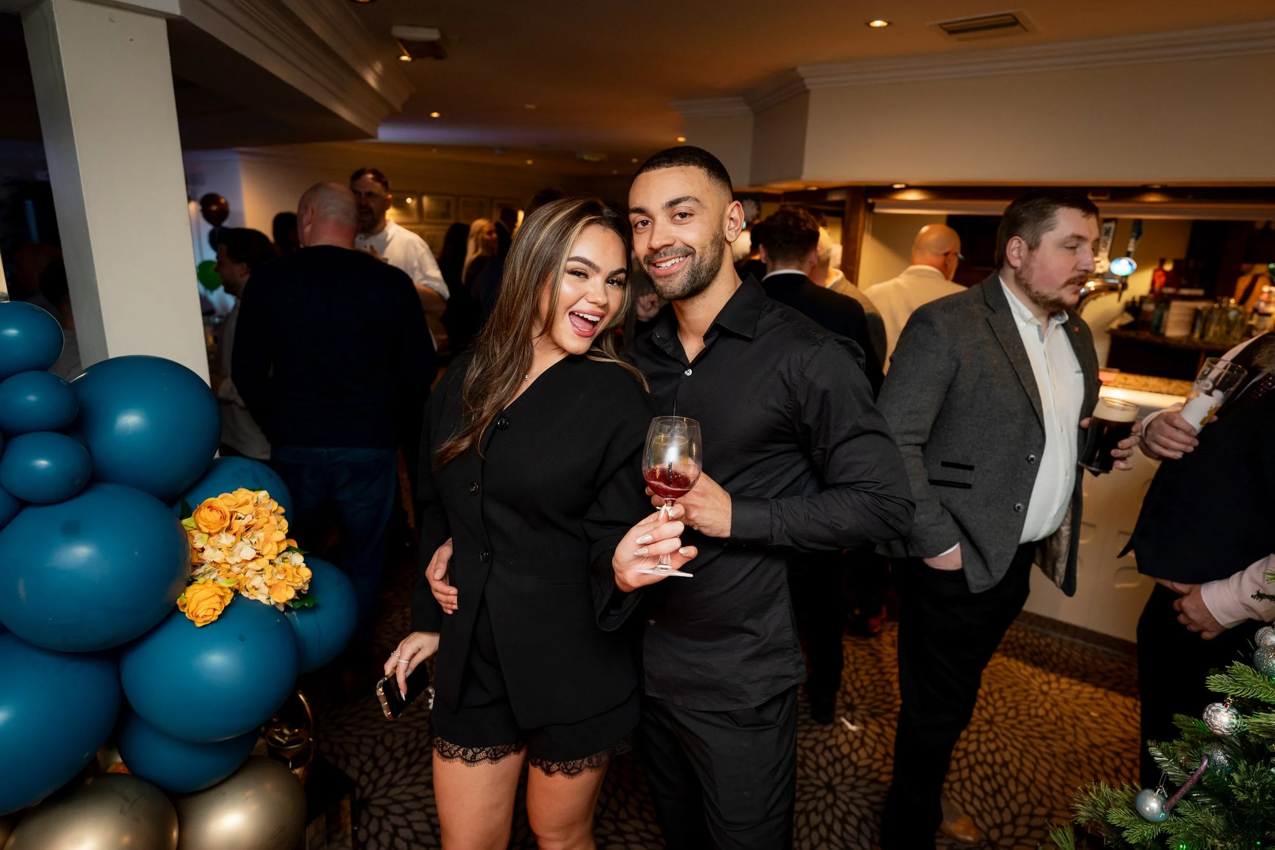 A woman and man smiling and holding drinks at a party, with other guests in the background and Christmas decorations nearby.