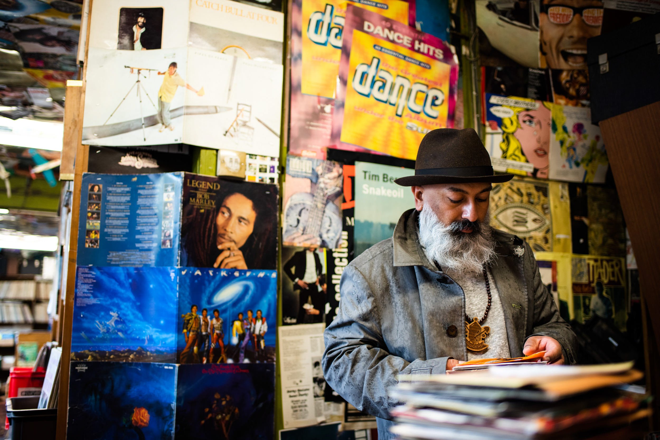 A man with a gray beard, wearing a black hat and a gray jacket, stands in front of a wall covered with colorful posters and album covers, looking down at a stack of records or magazines.