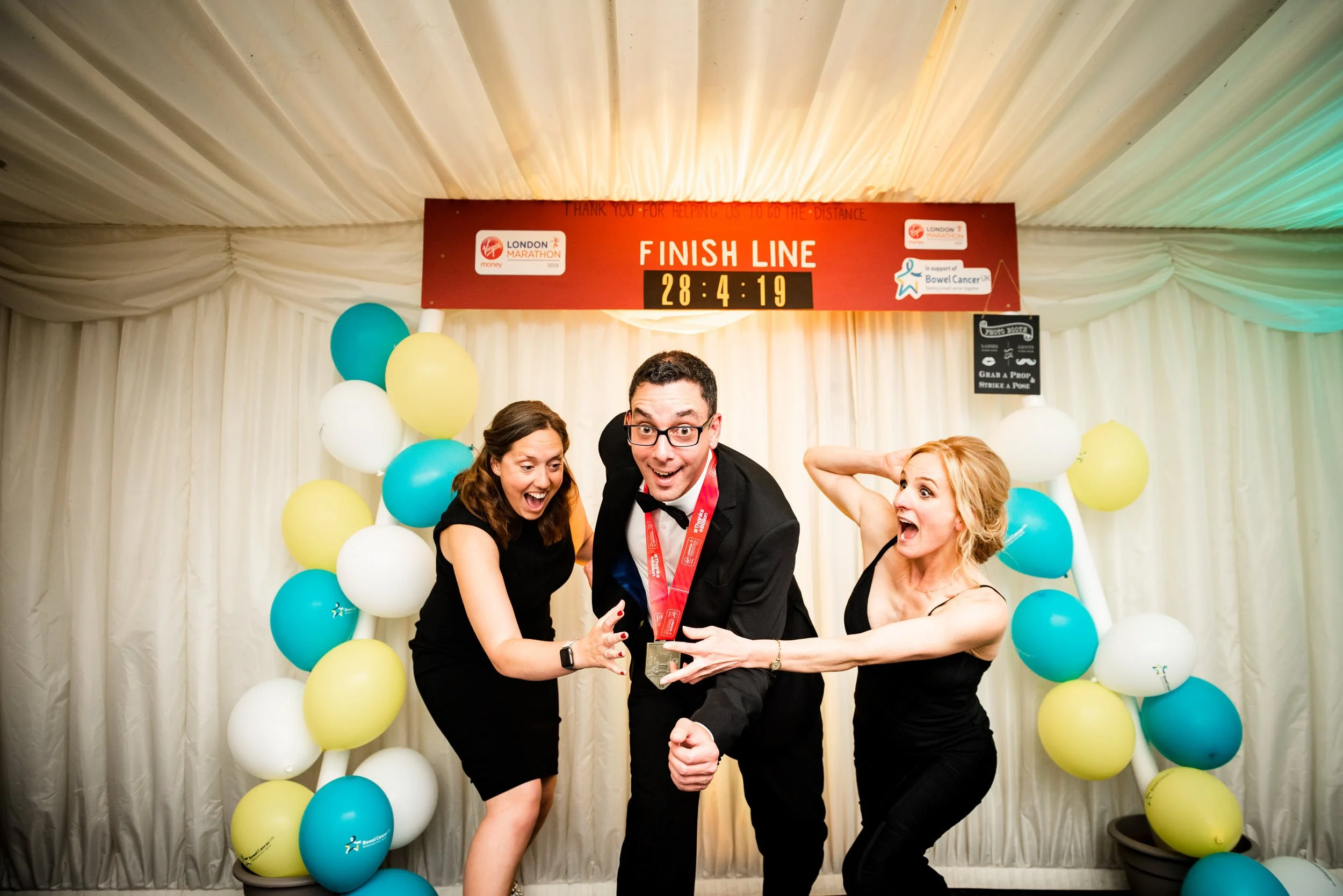 Three celebration participants in black attire at the finish line of a marathon, hugging and cheering as they celebrate their achievement. The backdrop features balloons in white, yellow, and teal, with a red finish line banner above showing the rema