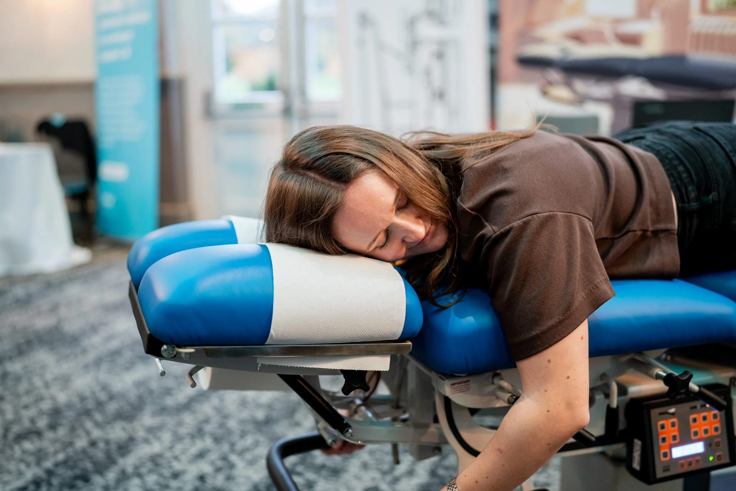 A woman lying face down on a modern medical or massage table with blue and white padding, resting her head on the padded headrest, at a professional conference for chiropractors