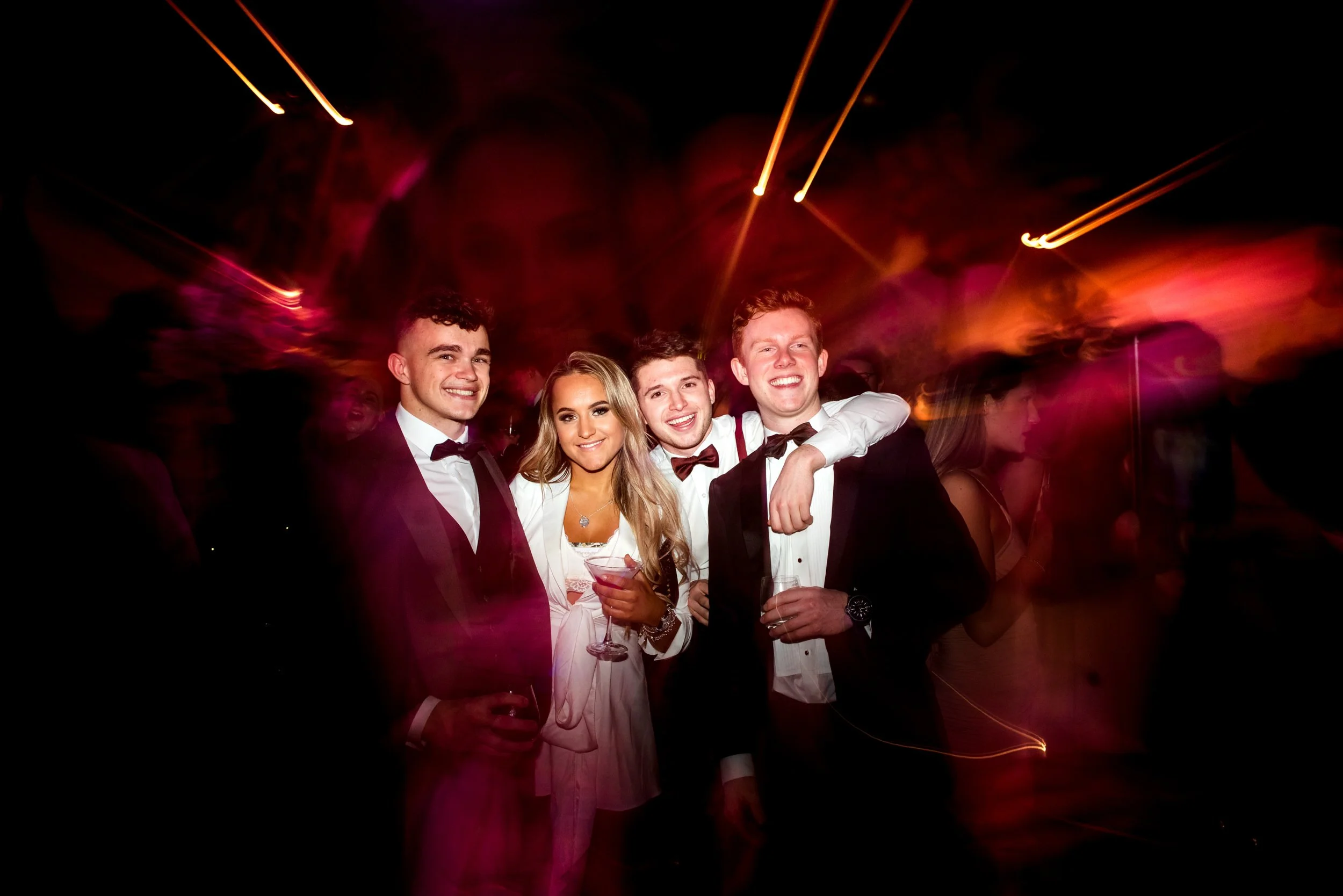 Group of four young adults dressed in formal attire at an evening reception with colorful blurred lights in the background.