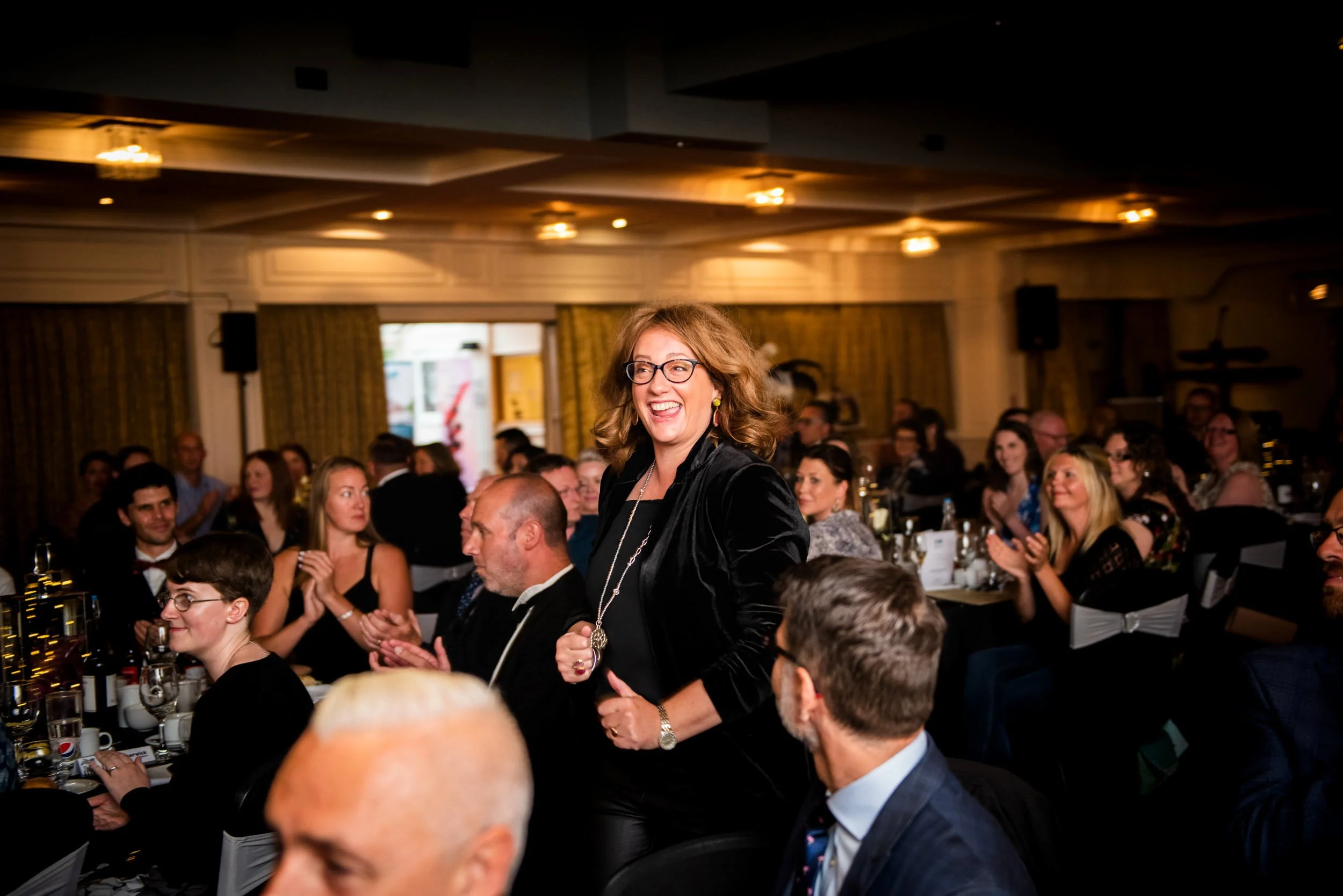 A woman with short curly hair, glasses, wearing a black velvet jacket, smiling confidently in a formal event, surrounded by seated guests at a banquet.