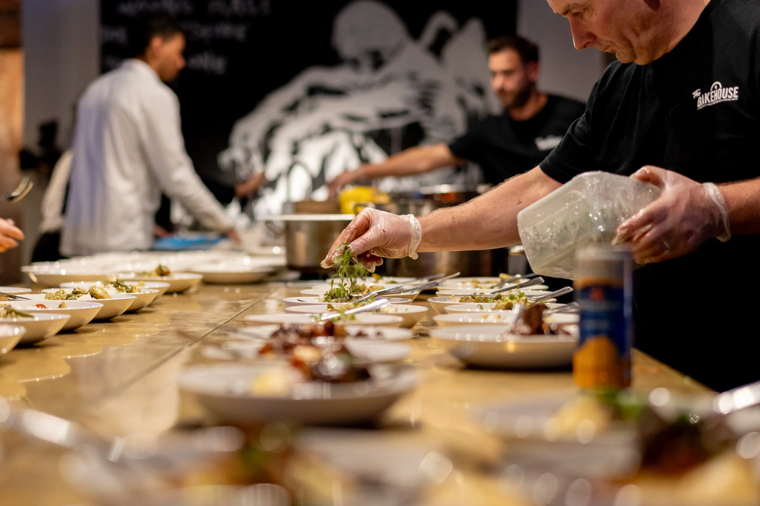A chef in black uniform and gloves garnishing plated dishes on a kitchen counter with other staff in the background.