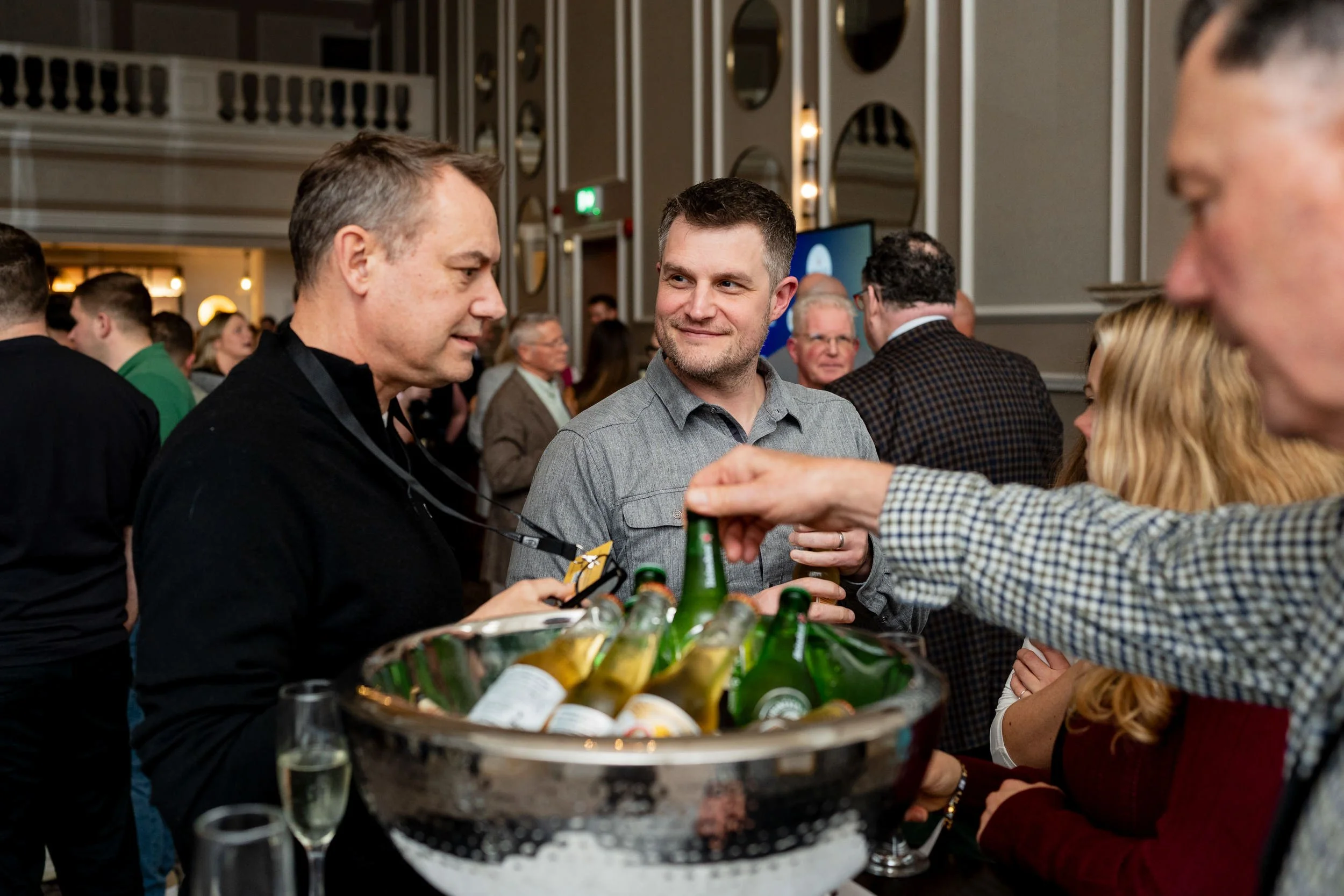 People socializing at an evening party, with a man serving drinks from a tray of bottled beers.