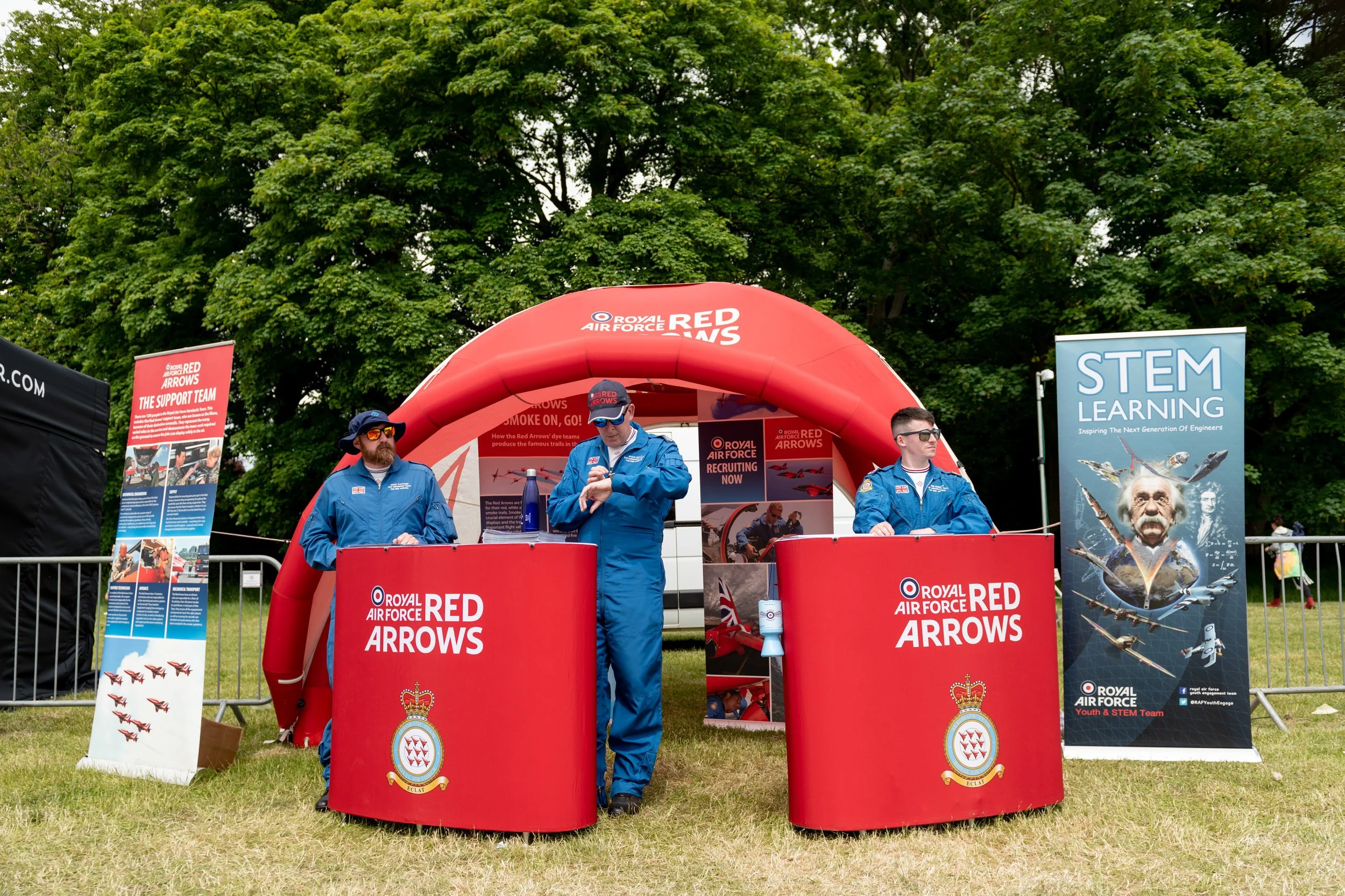 Three men in blue uniforms standing behind red booths with 'Royal Air Force Red Arrows' branding at the Midlands Air Festival . Behind them is a red inflatable arch, and there are informational signs about the Red Arrows.
