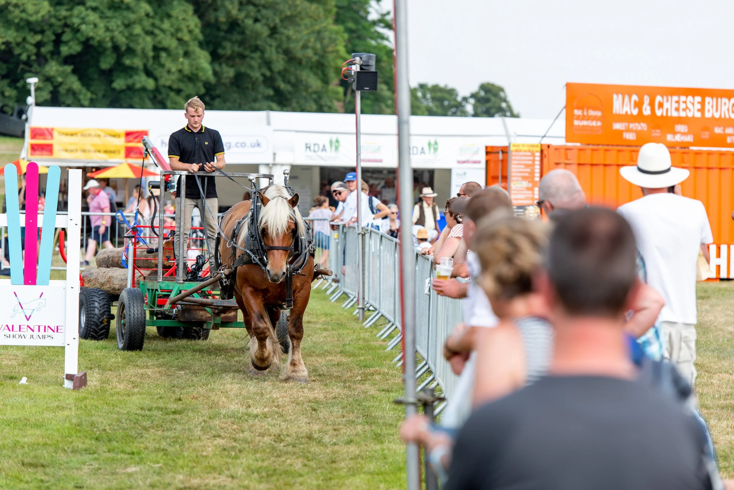 A horse pulling a cart with logs at the Midlands Game Fair, with spectators watching behind a fence and food stands in the background.