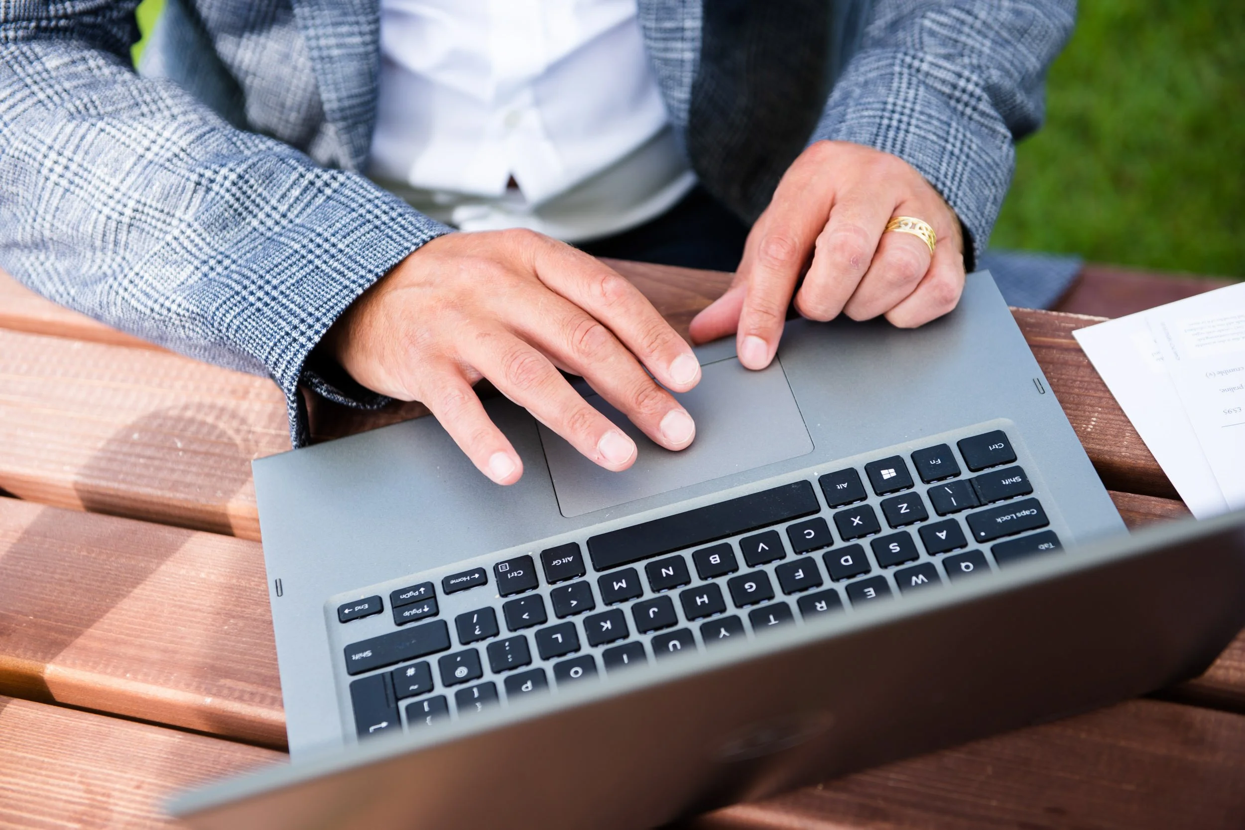Person in gray plaid blazer typing on a silver laptop on a wooden table outdoors
