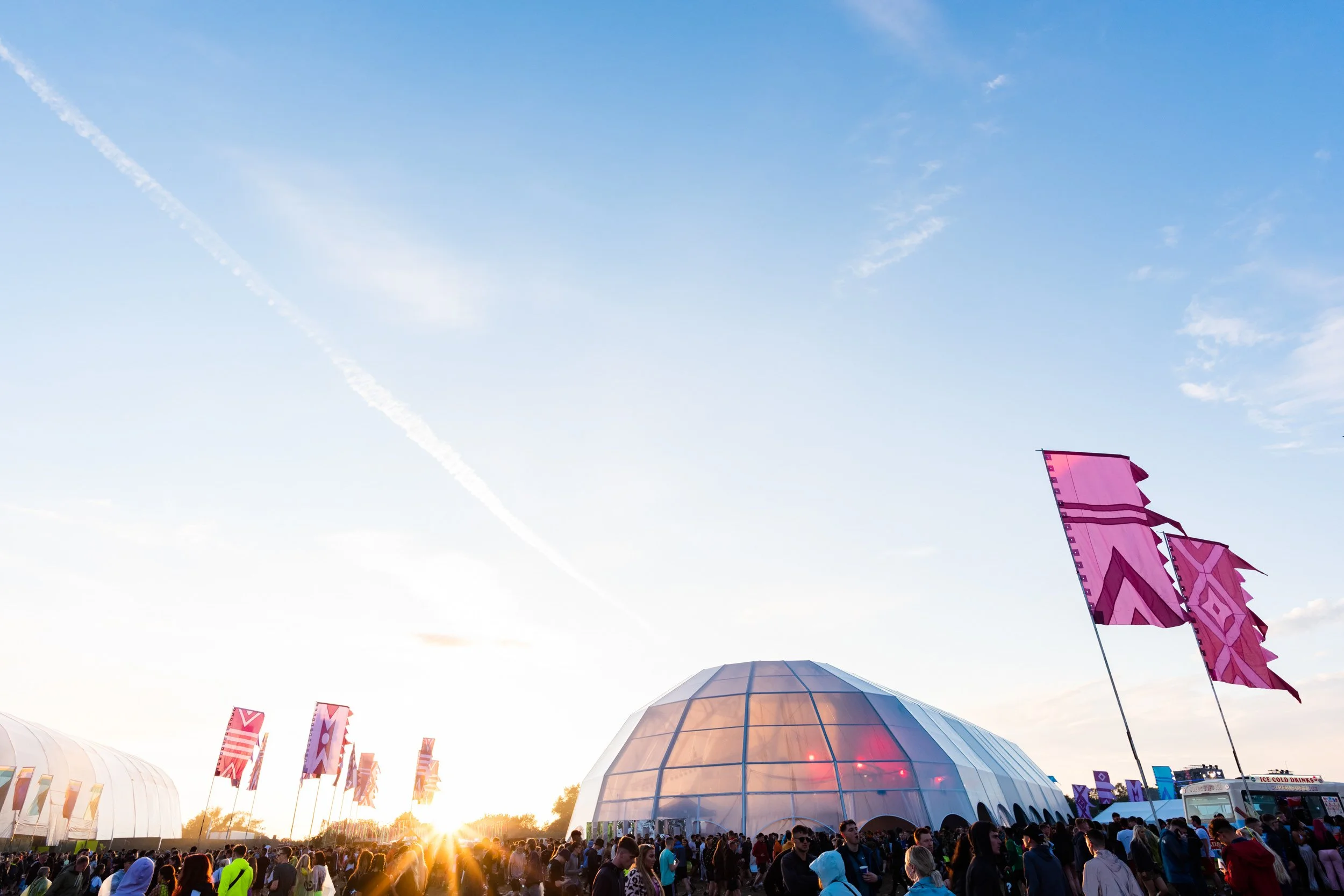 A large crowd of people gathering outside at the outdoor festival ParkLife during sunset, with a geodesic dome structure by Fews Marquees and pink flags in the background.