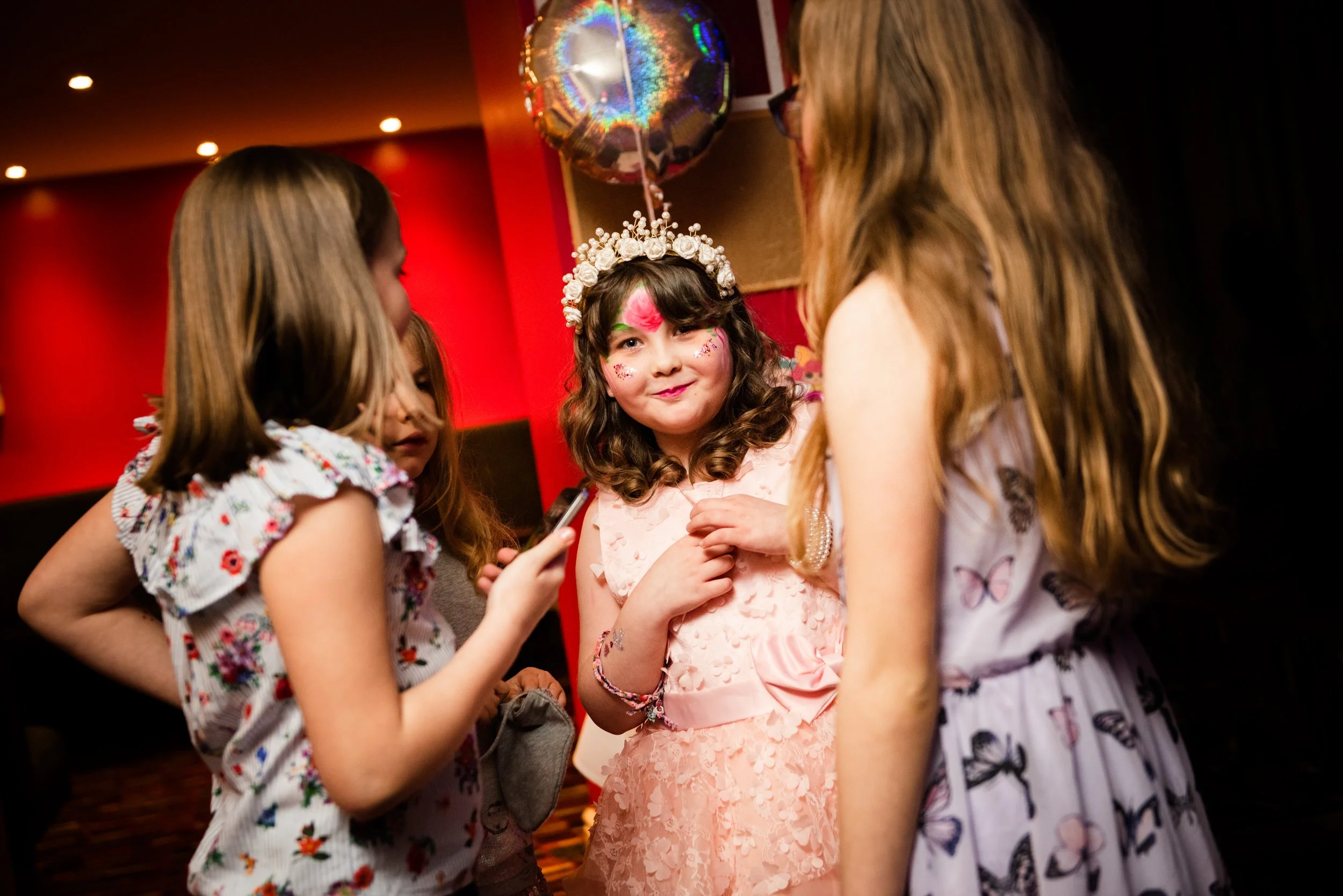 A birthday girl with face paint and a floral headband stands in the center of a group of girls at a birthday party, with colorful decorations and balloons in the background.