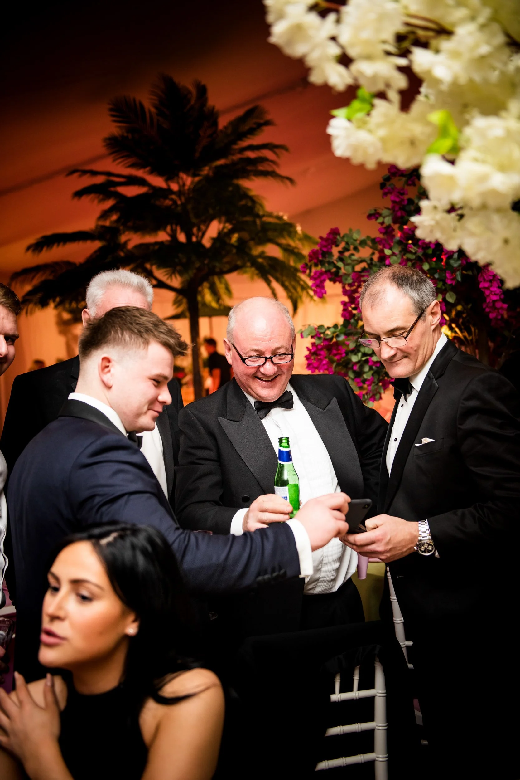 Group of people dressed in formal tuxedos at a festive event, looking at a smartphone, with a woman in the foreground and tropical plants in the background.