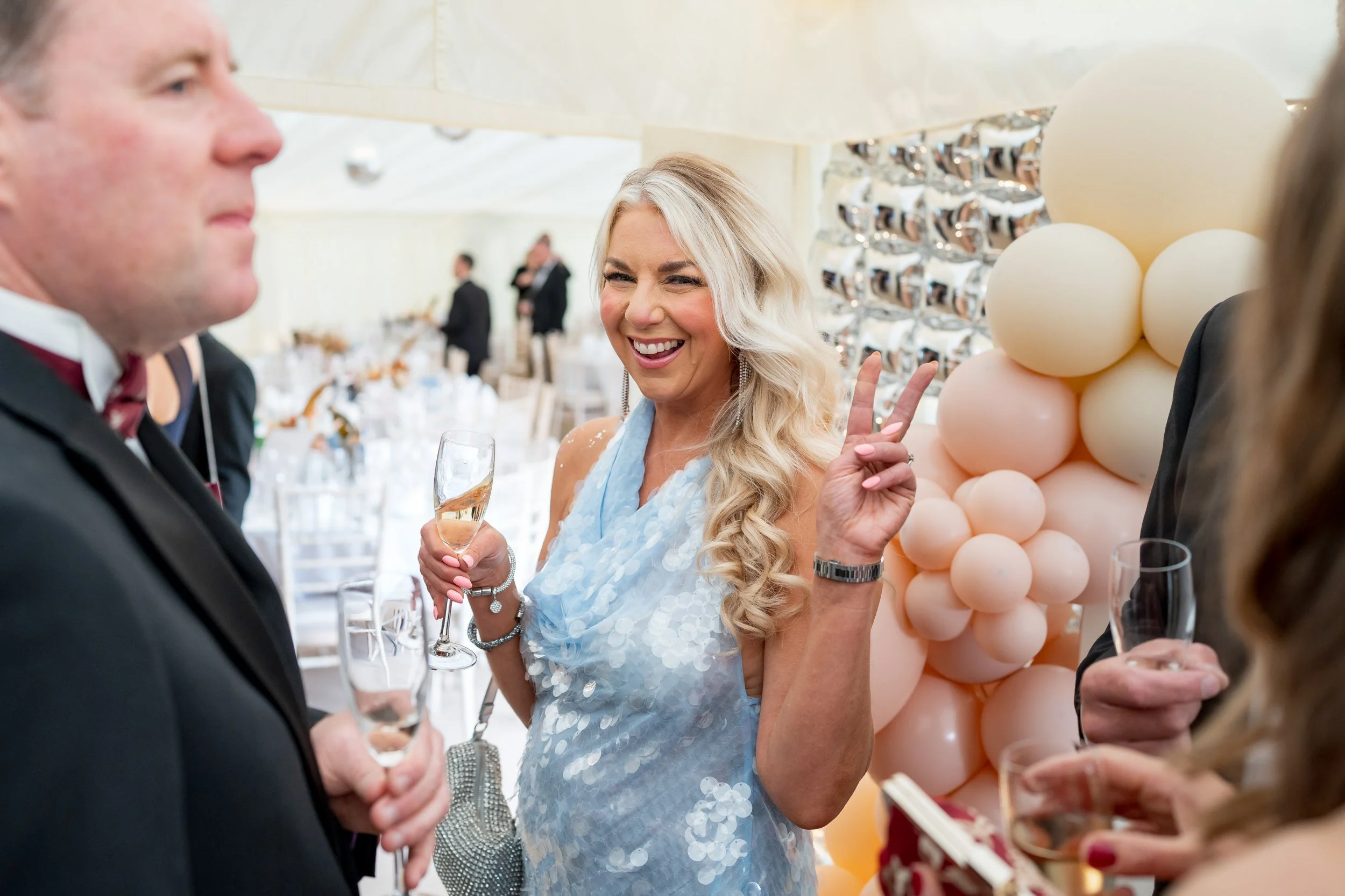 A woman with long blonde hair in a light blue dress making a peace sign with her fingers and smiling at a social event with people holding glasses of champagne and a balloon decoration in the background.