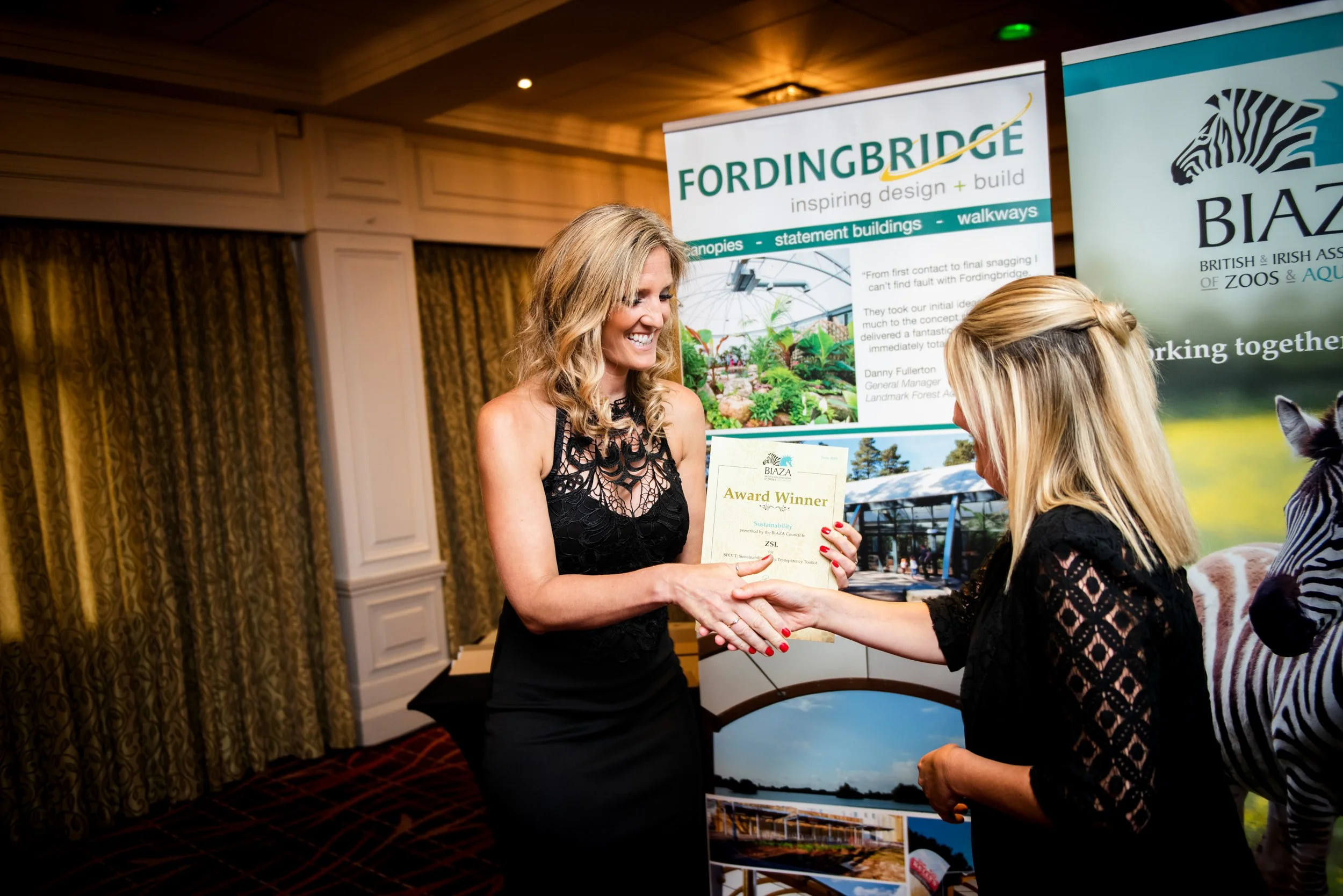 A woman in a black dress smiling and holding an award certificate is shaking hands with another woman at an indoor event. There are promotional banners in the background, one for Fordingbridge and another for Biaza, showing images of parks and animal