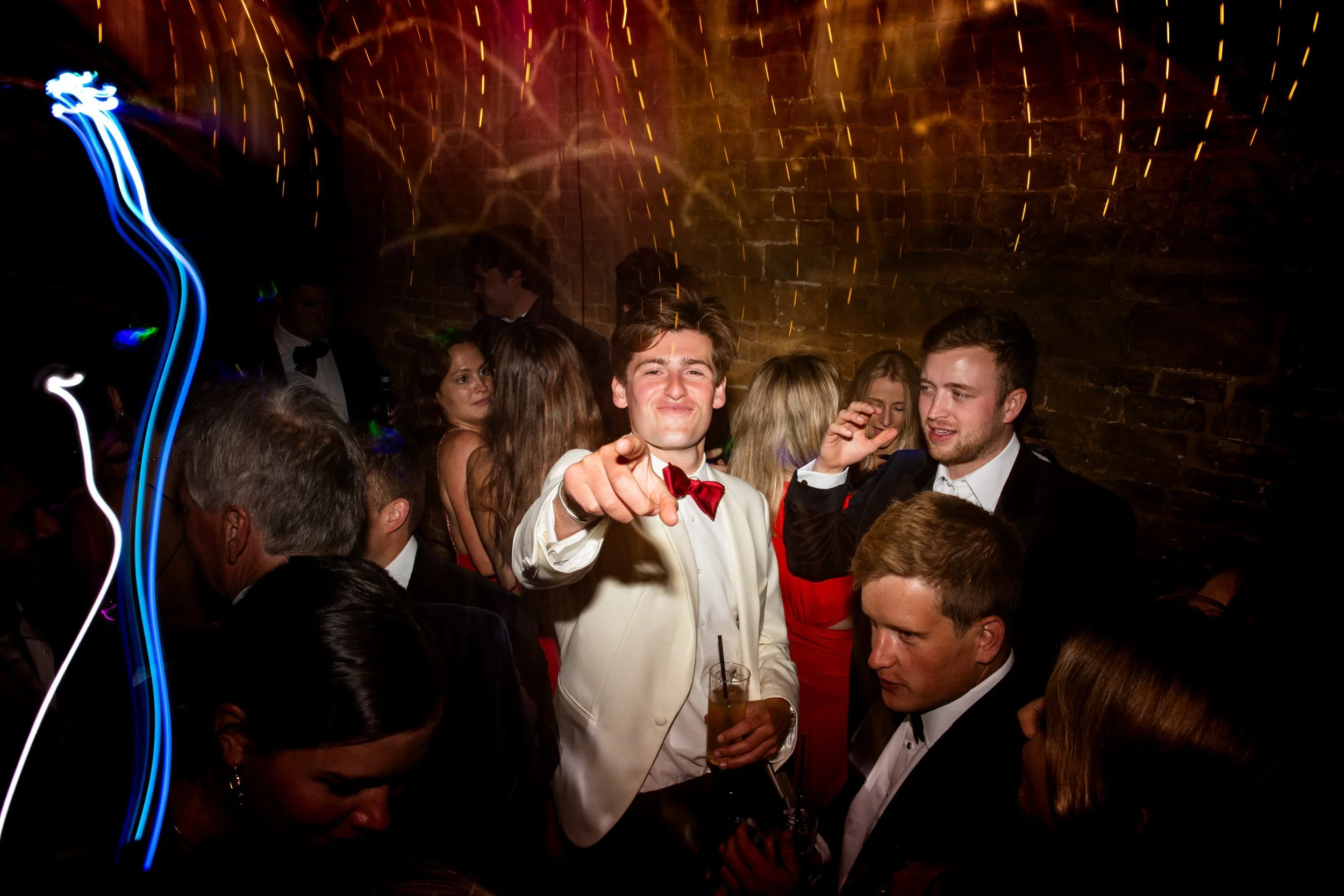 Group of people in formal attire dancing and socializing at a celebration party with colorful lights and a brick wall background.