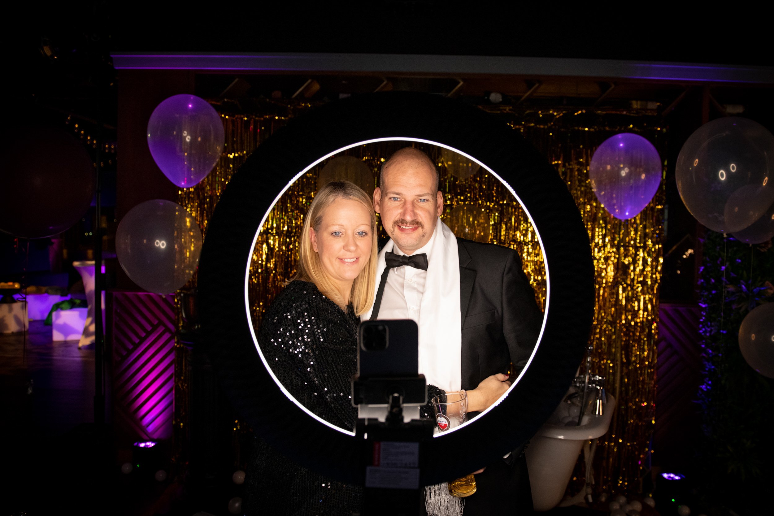 A couple dressed in formal attire taking a selfie through a circular LED light frame at a PS Events party with golden decorations, balloons, and purple lighting.