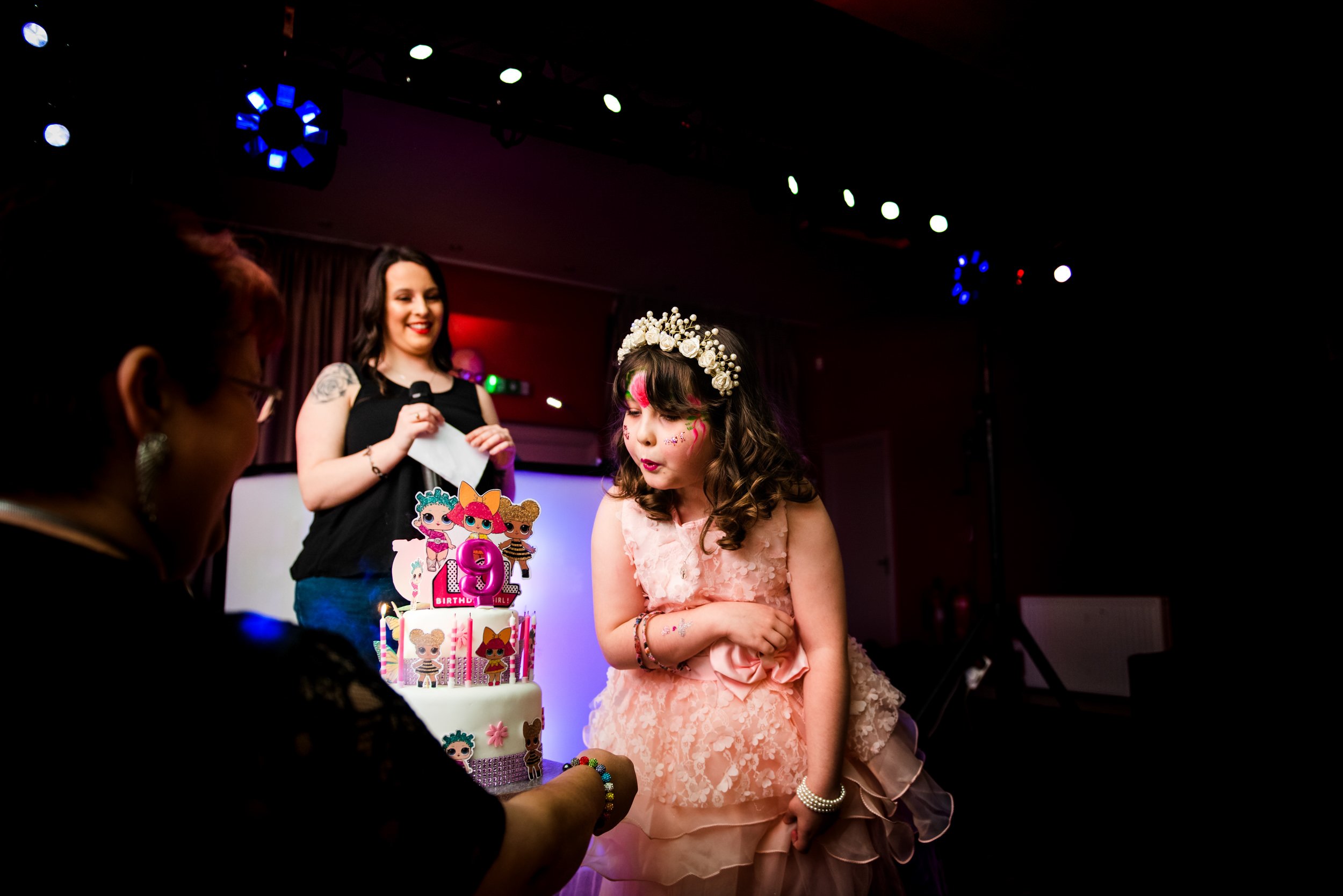 A young girl in a peach dress with face paint and a floral headband blowing out candles on a birthday cake at her celebration, with two women standing nearby in a dimly lit room with celebration decorations.