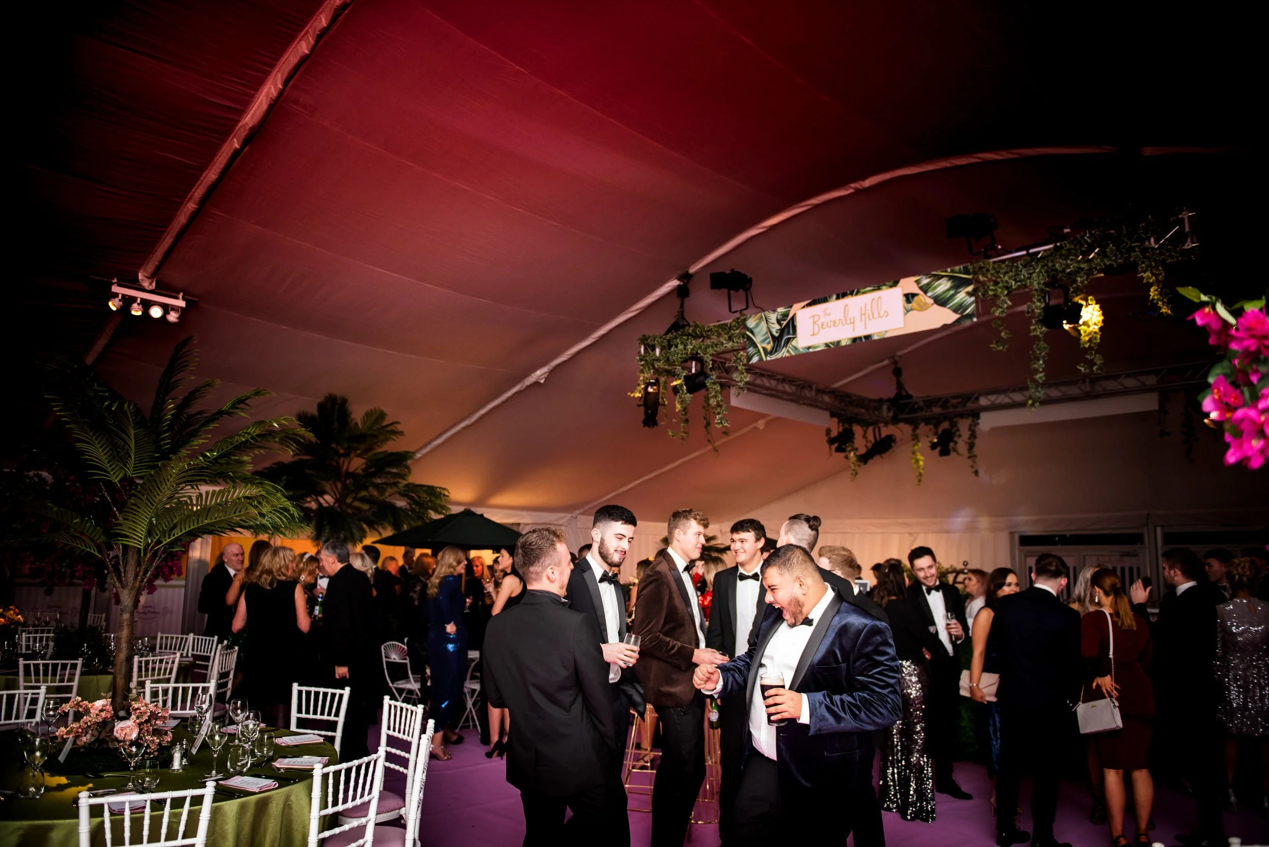 People dressed in formal attire, socializing and enjoying drinks at a party inside a decorated tent with tropical plants and a sign that says "Beverly Hills."