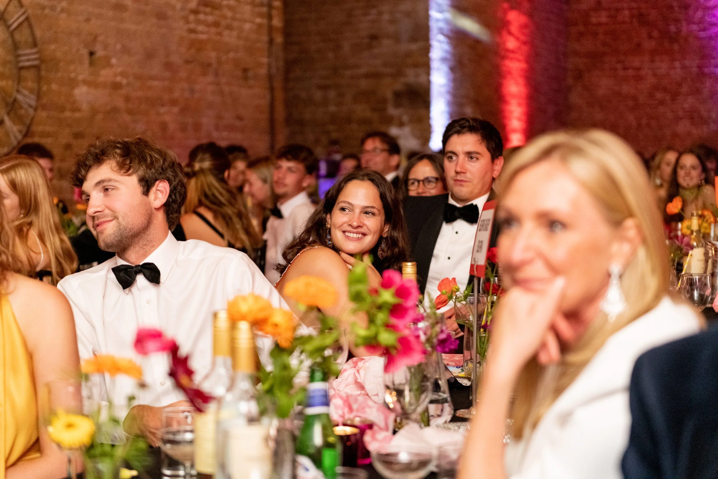 People attending a formal event or dinner, dressed in tuxedos and evening gowns, seated at tables with colorful flowers and bottles, inside an industrial brick-walled venue.
