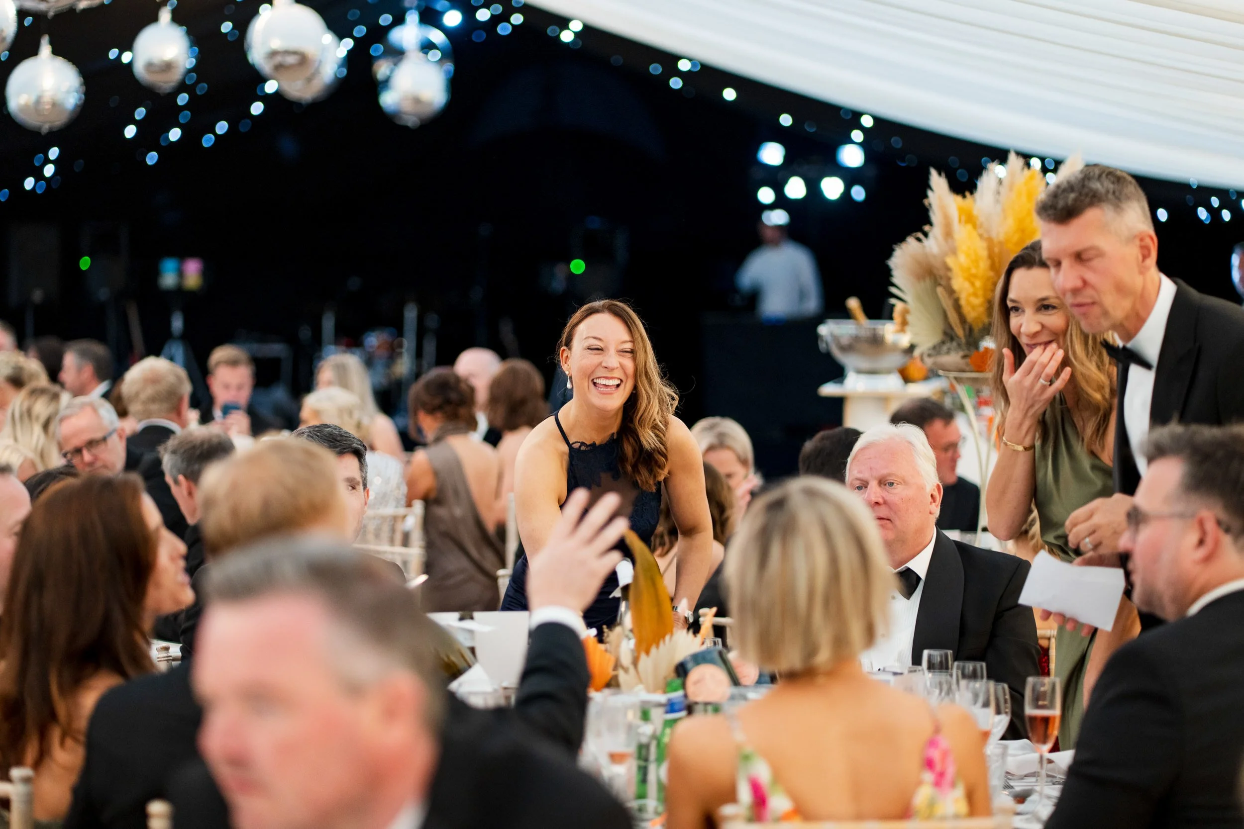 People at a formal event or banquet, some are smiling and engaging in conversation, with a woman standing and laughing in the center, and decore, including balloons and floral arrangements, in the background.