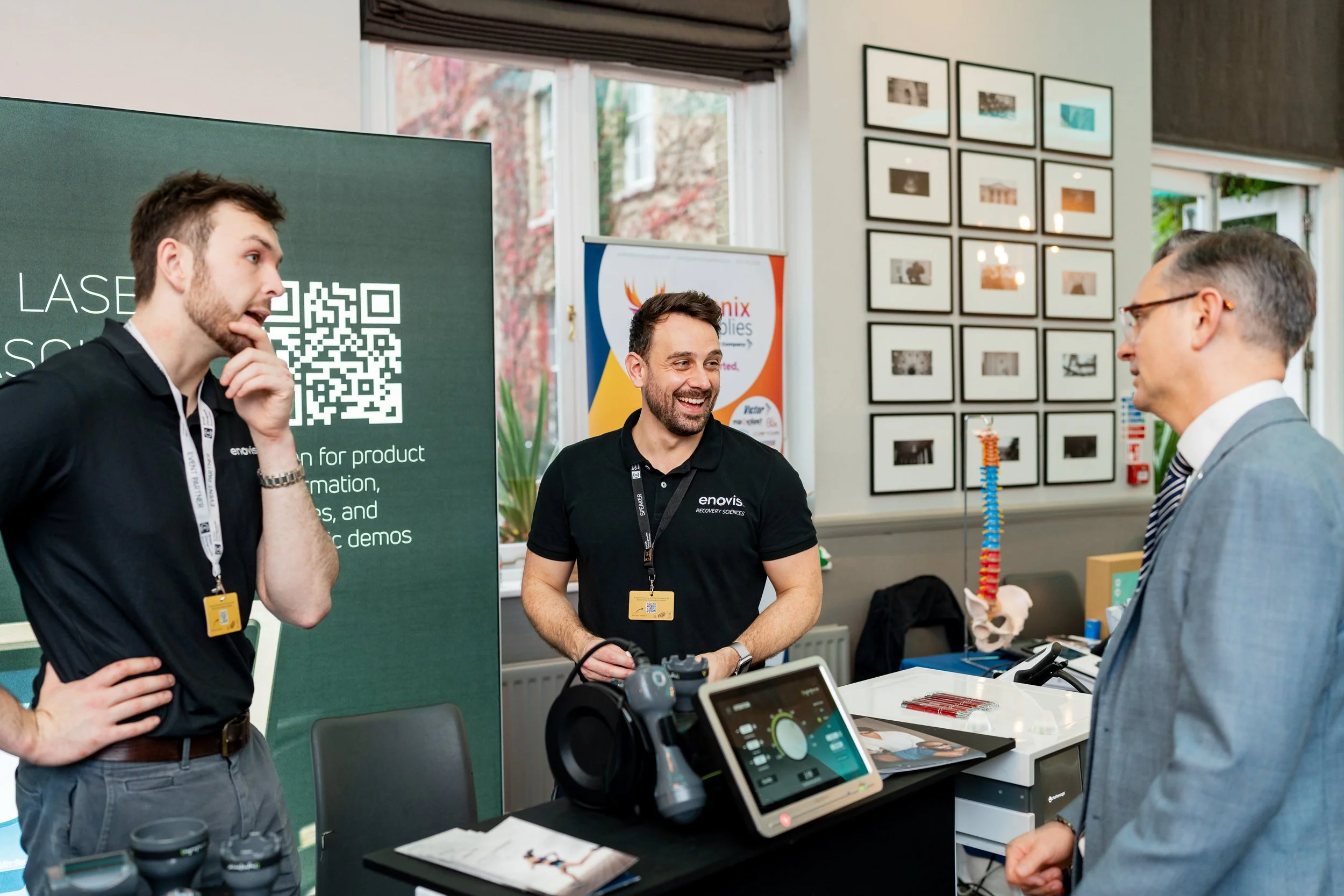 Three men at a trade show booth engaging in conversation. Two are wearing black shirts with 'enovis' logos, and the third man is dressed in a gray suit. The booth features a green display board with a QR code and text, a monitor, and scientific model