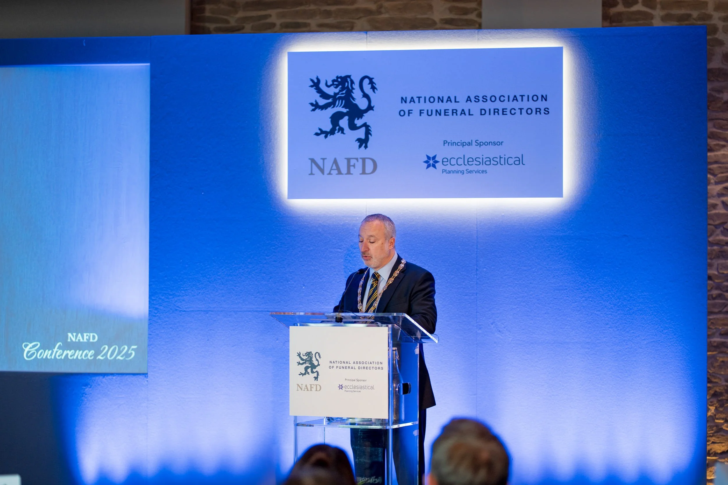 A man in a suit with a patterned tie and a chain of office standing at a podium on a stage with a blue background, during the NAFD Conference 2025, with a screen displaying the NAFD logo, a lion emblem, and the text 'National Association of Funeral D