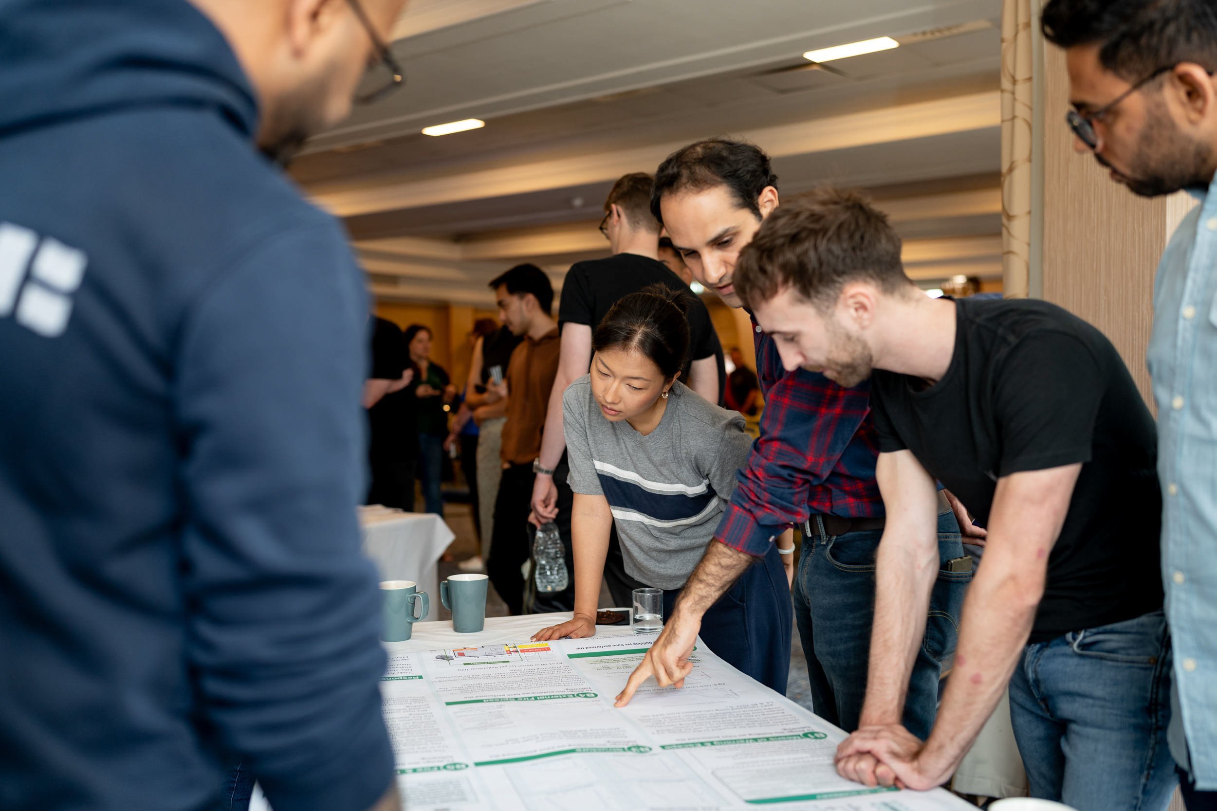 Group of people gathered around a table examining printed documents, with coffee mugs and a water bottle present, in a busy indoor setting.