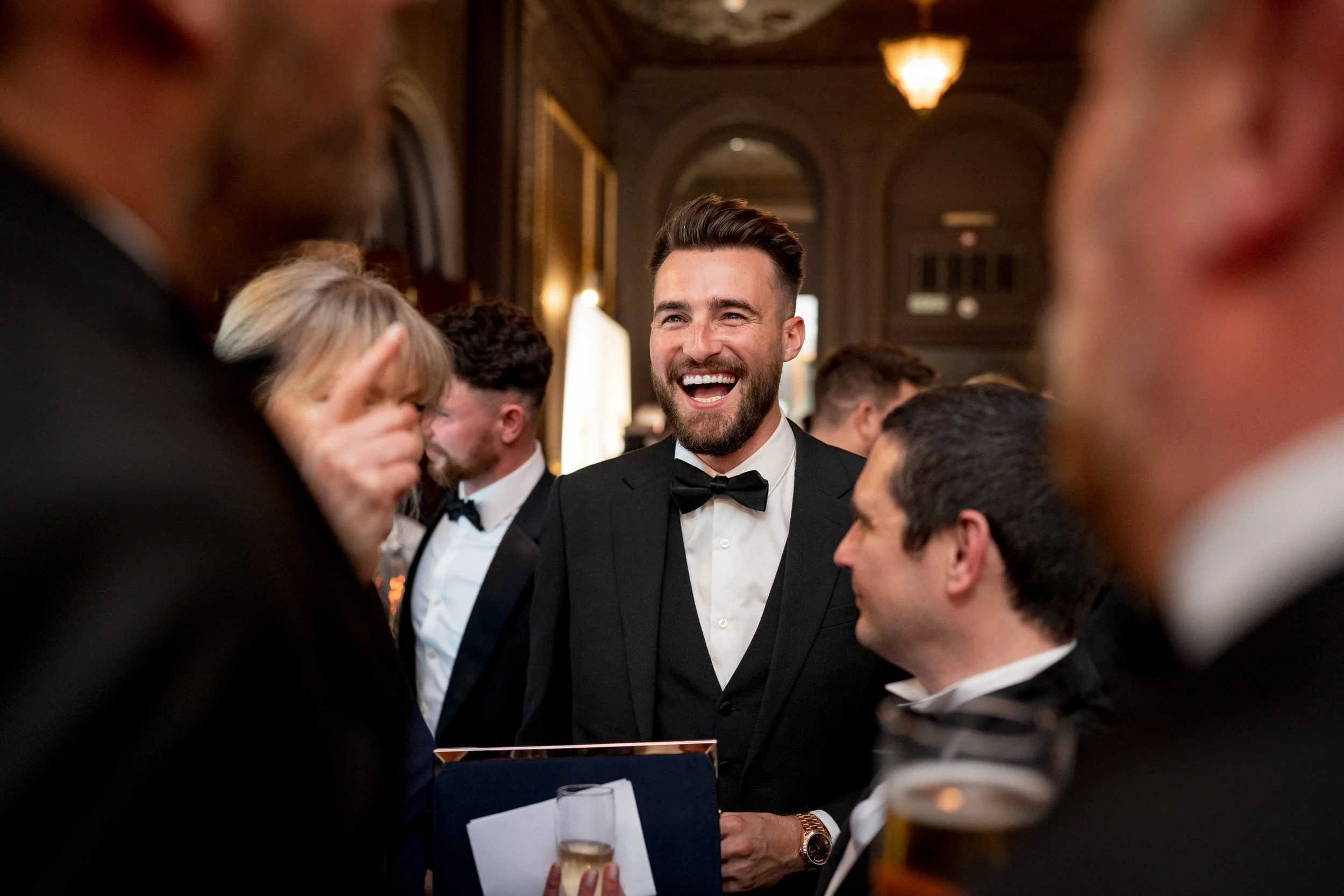 A group of people dressed in formal tuxedos and evening gowns at a celebration or event, with a man in the center smiling and laughing.