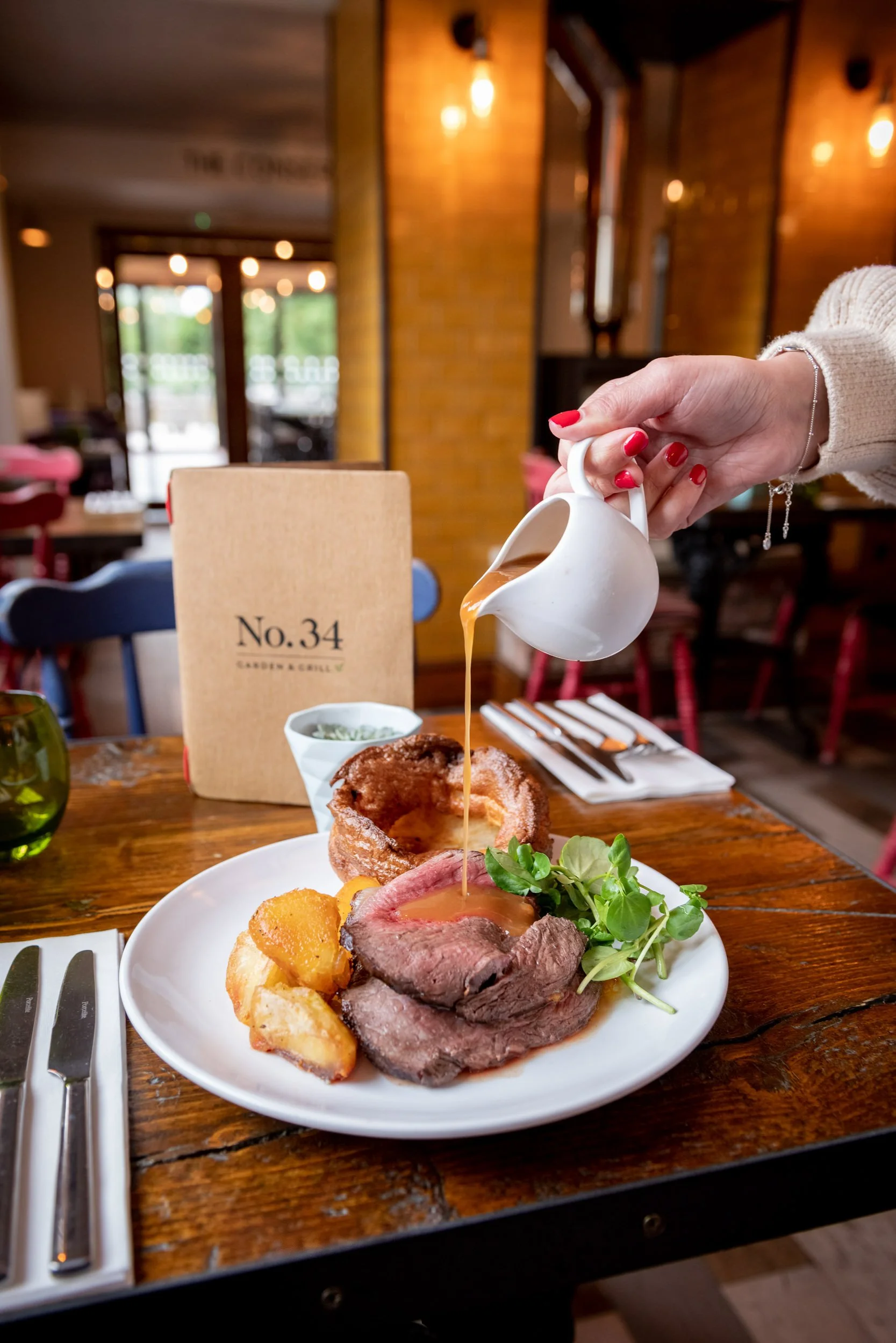 A person pouring gravy over a plate with roast beef, roast potatoes, Yorkshire pudding, and greens, in a cozy restaurant setting.