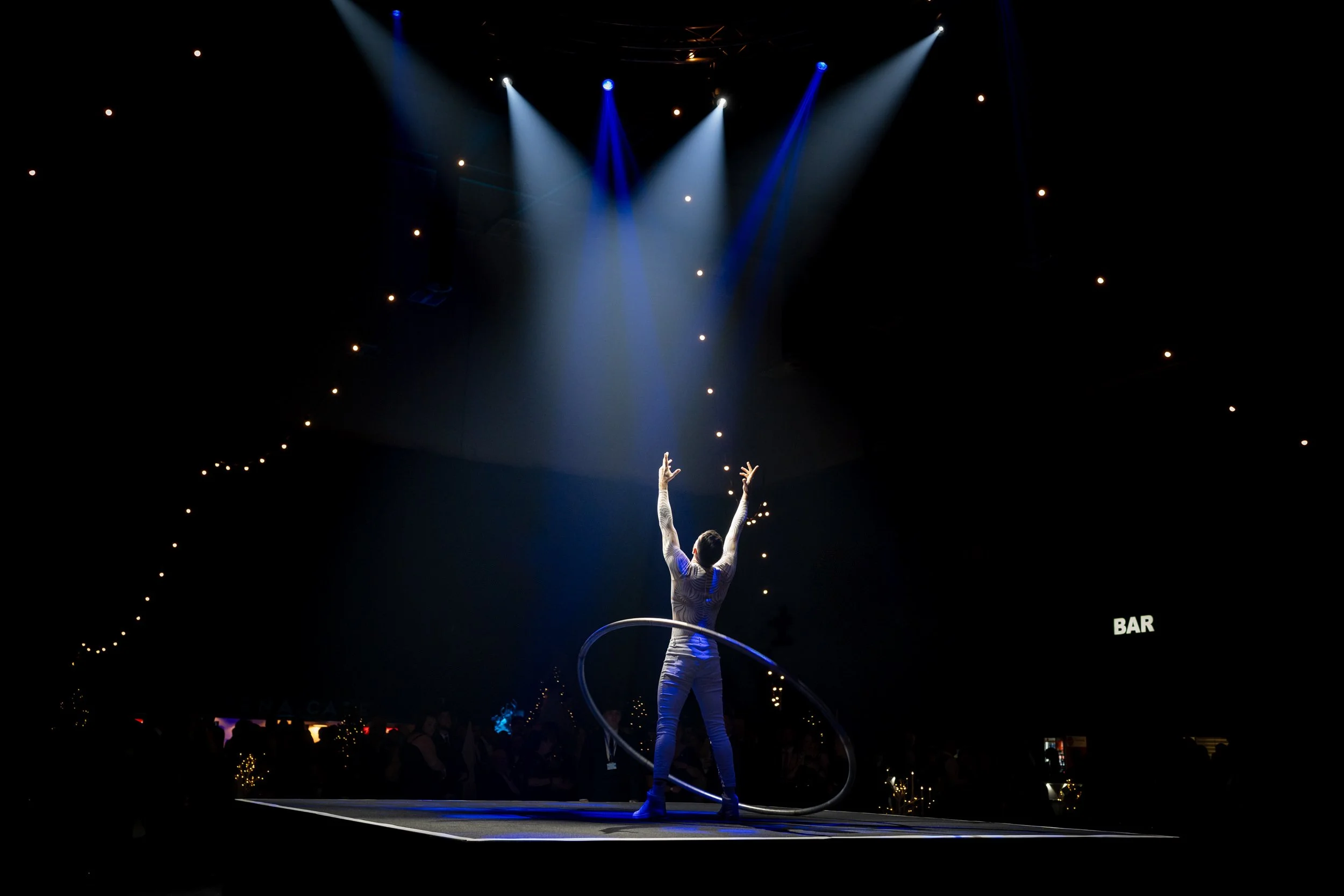 An acrobat performer on stage with arms raised, standing inside a hula hoop, illuminated by blue and white stage lights in a dark venue Hall for a luxury works Christmas Party with ceiling string lights and a visible bar sign in the background.