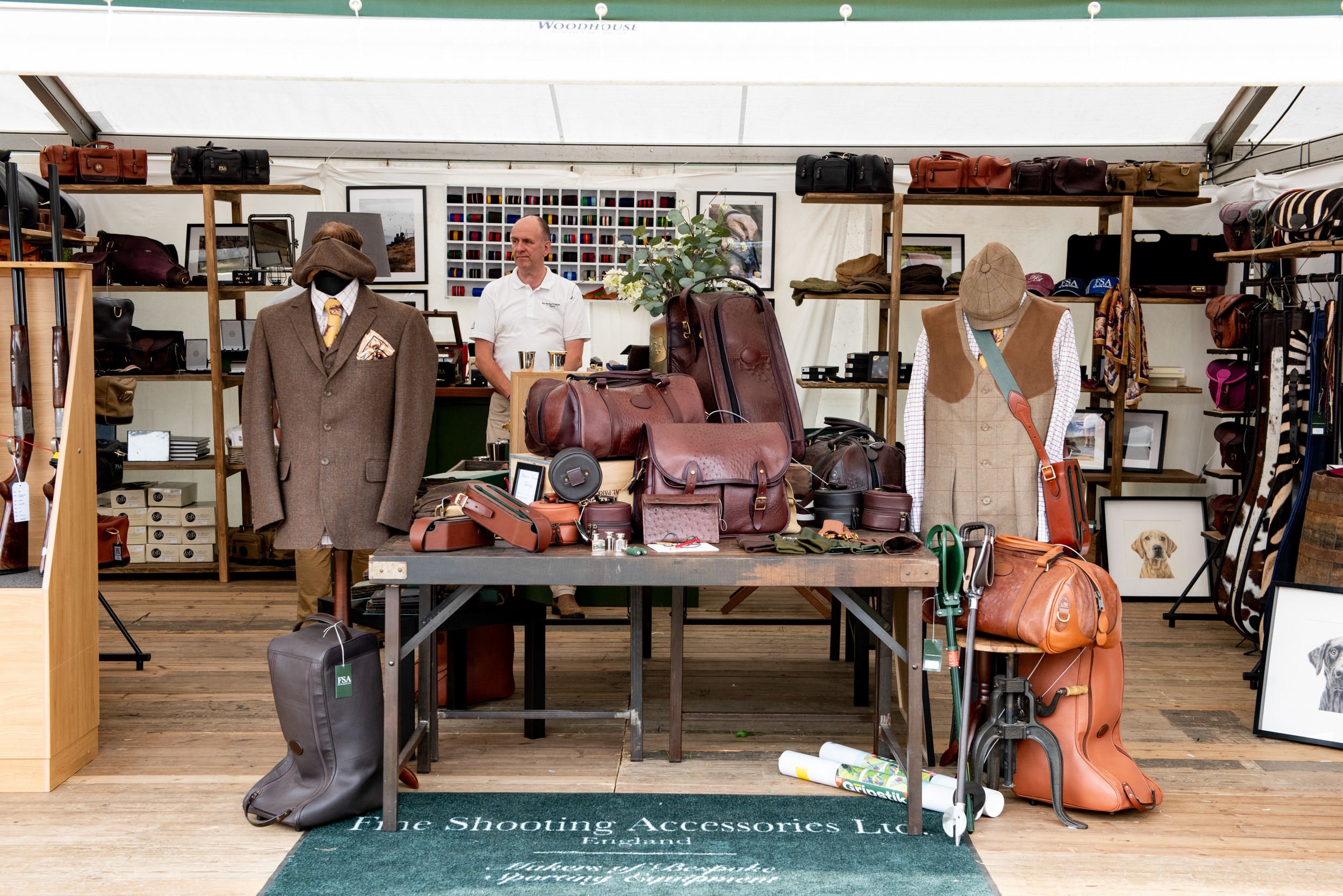 A booth displaying various leather and canvas shooting accessories and outdoor clothing, including suits, bags, and hats, with a person in a white shirt standing behind the display.