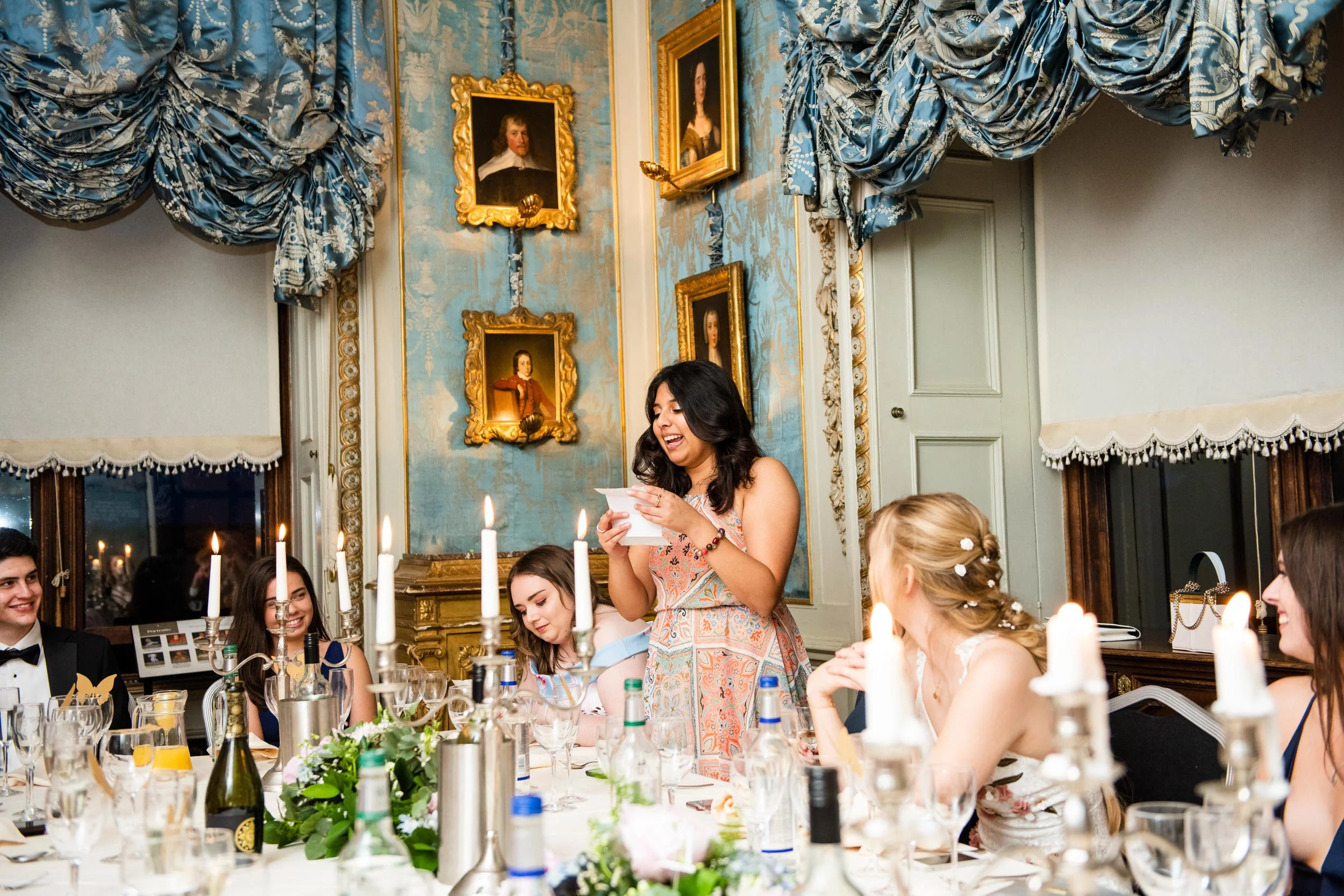 A woman giving a speech at a formal dinner party with guests seated around the table, decorated with candles, flowers, and bottles, in Warwick Castles elegant Queen Elizabeth room with ornate curtains and framed portraits on the wall.