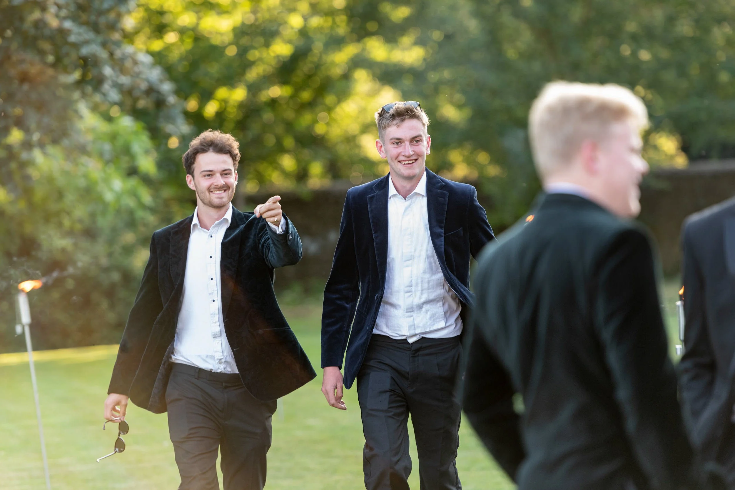 Three men in formal attire walking outdoors at a garden birthday party on a sunny day, smiling and enjoying themselves.