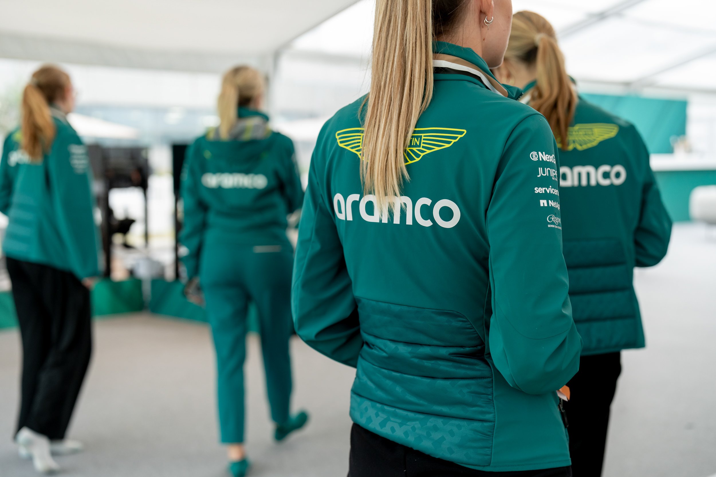 Women wearing teal jackets with 'aramco' logo and other sponsors, standing inside a tent or pavilion.